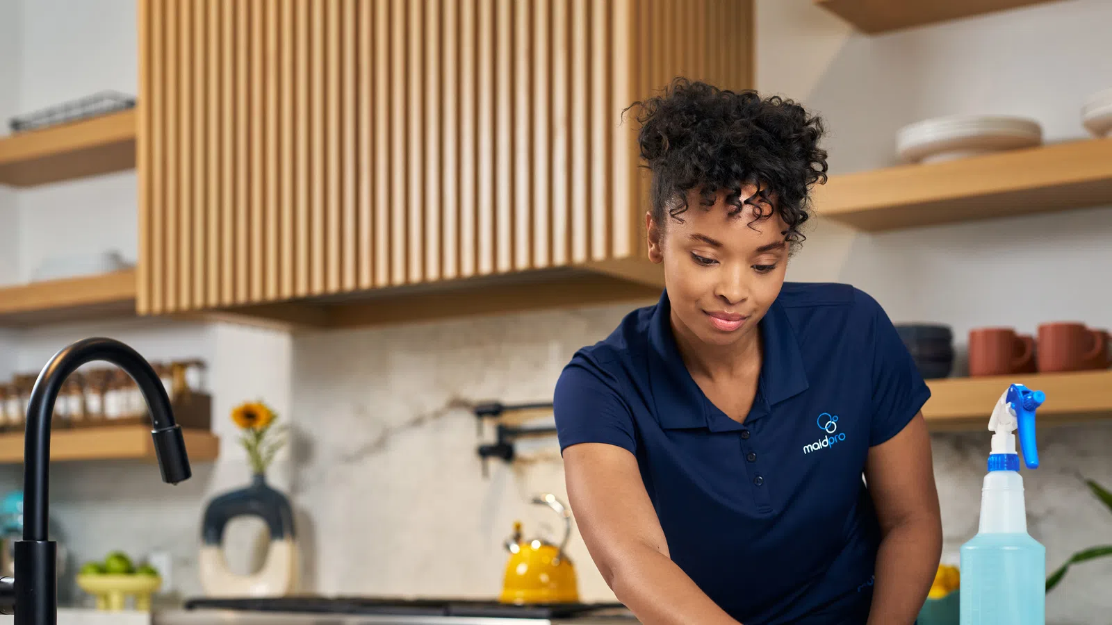 Woman in blue gloves cleaning kitchen countertop with spray bottle and cloth in modern kitchen.