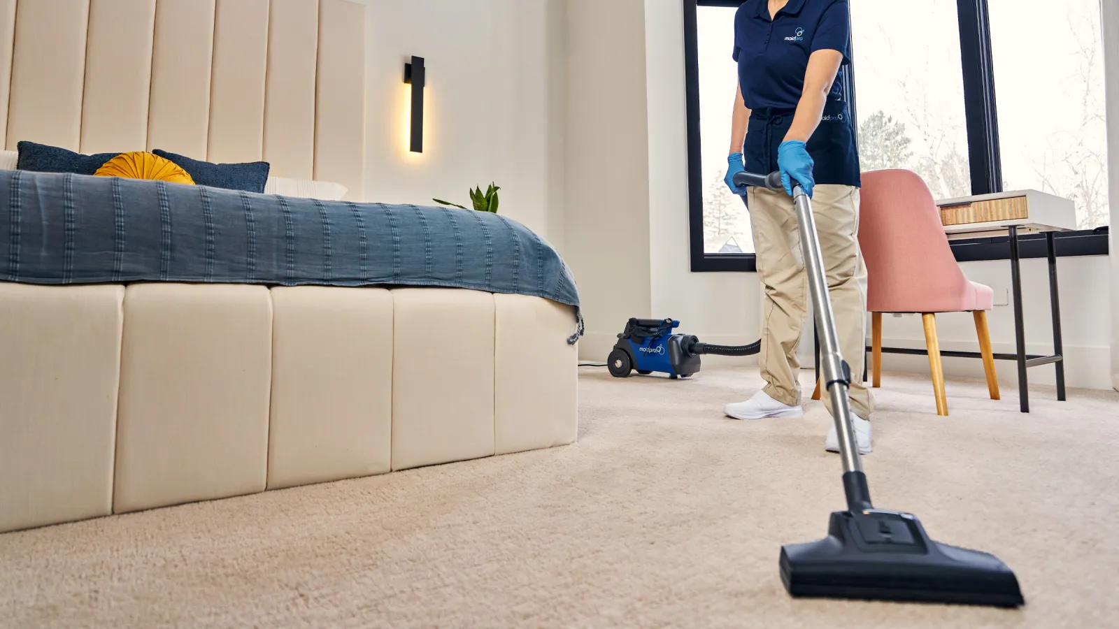 Professional cleaner vacuuming carpet in a modern bedroom with large windows and beige decor.