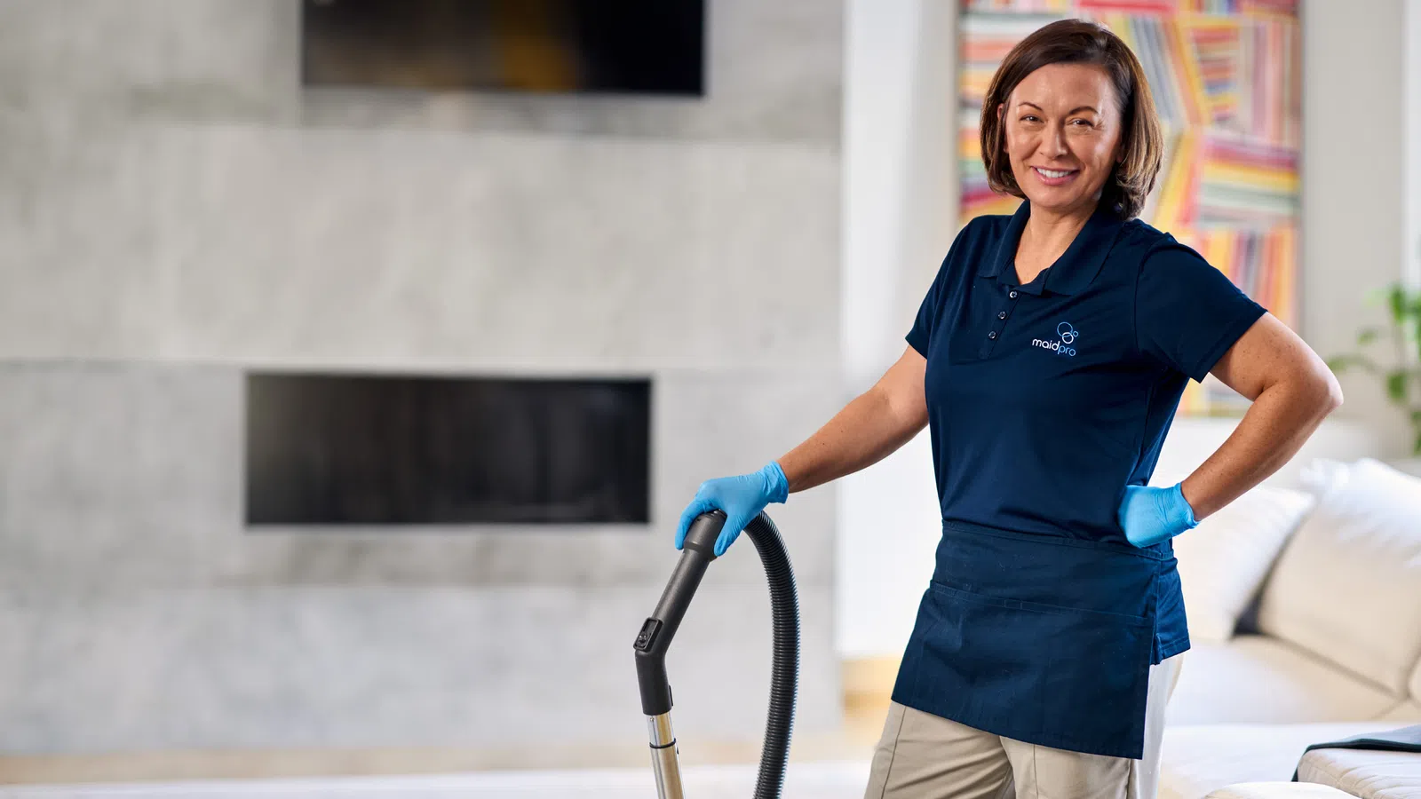 Smiling woman in uniform with gloves holding vacuum cleaner in a modern living room next to a white couch.