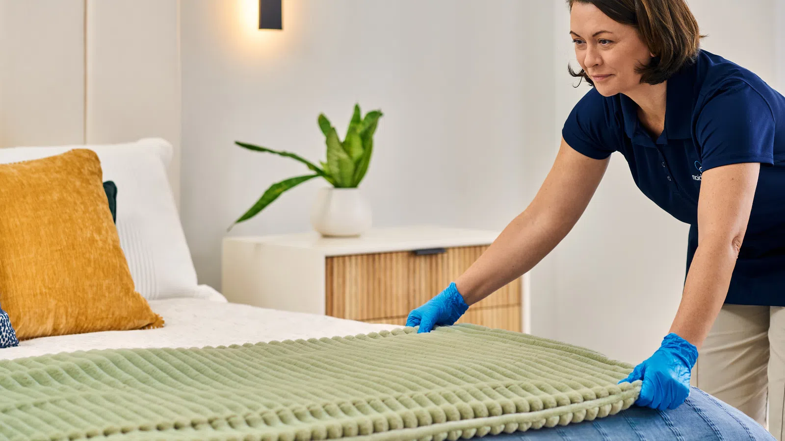 Housekeeper wearing gloves making a bed with green blanket in a bright bedroom with modern decor.