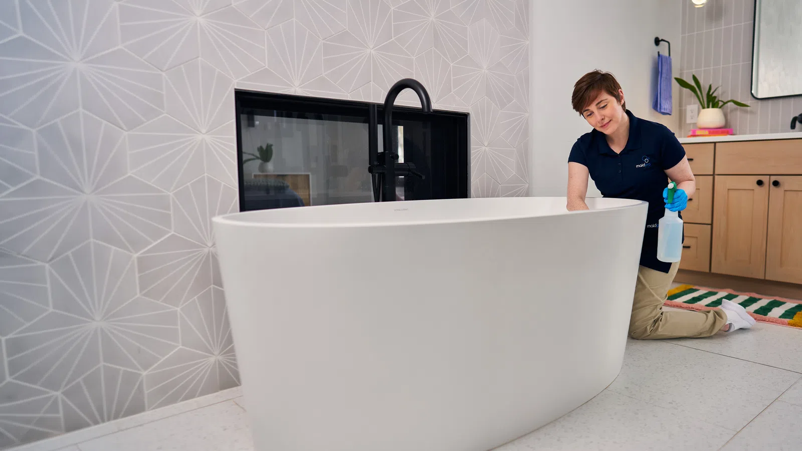 Person kneeling and cleaning a modern white freestanding bathtub in a stylish bathroom with geometric tiled walls.