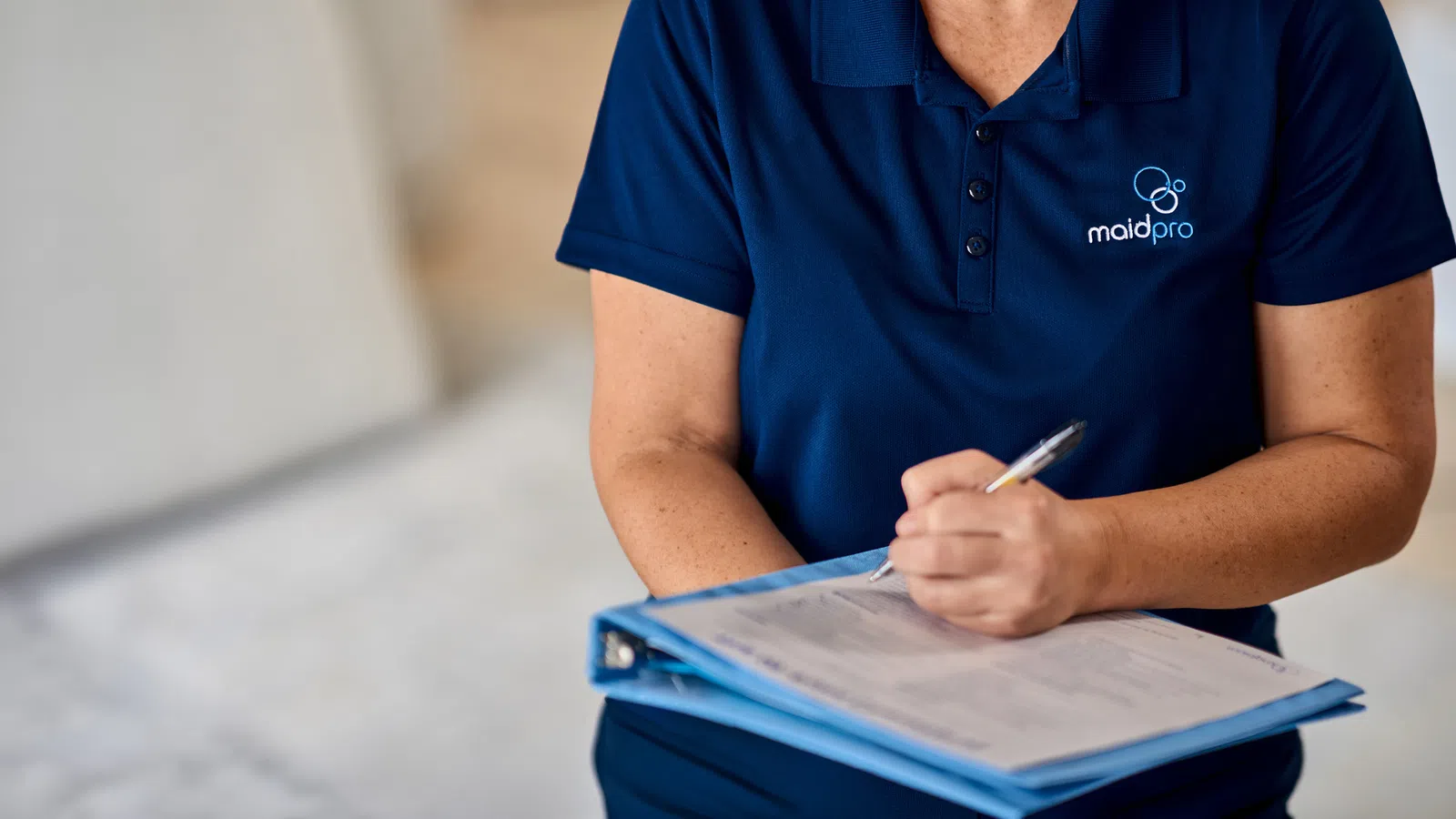 Professional cleaner in a navy MaidPro uniform filling out a checklist on a clipboard in a bright room.