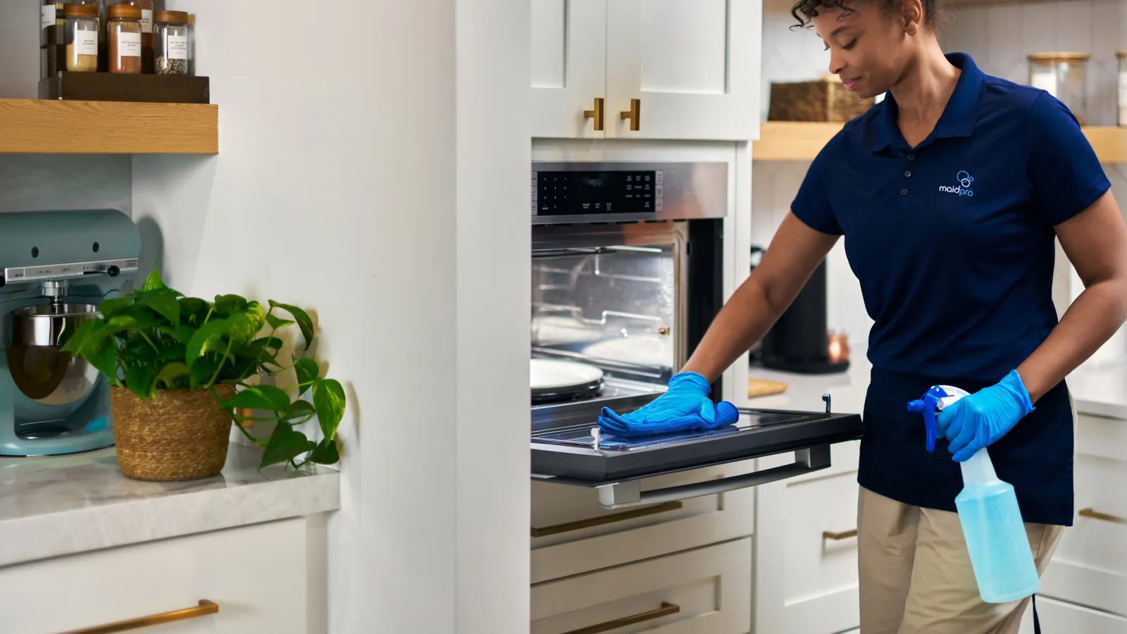 Professional cleaner in blue gloves wiping oven door with spray bottle in a bright modern kitchen.