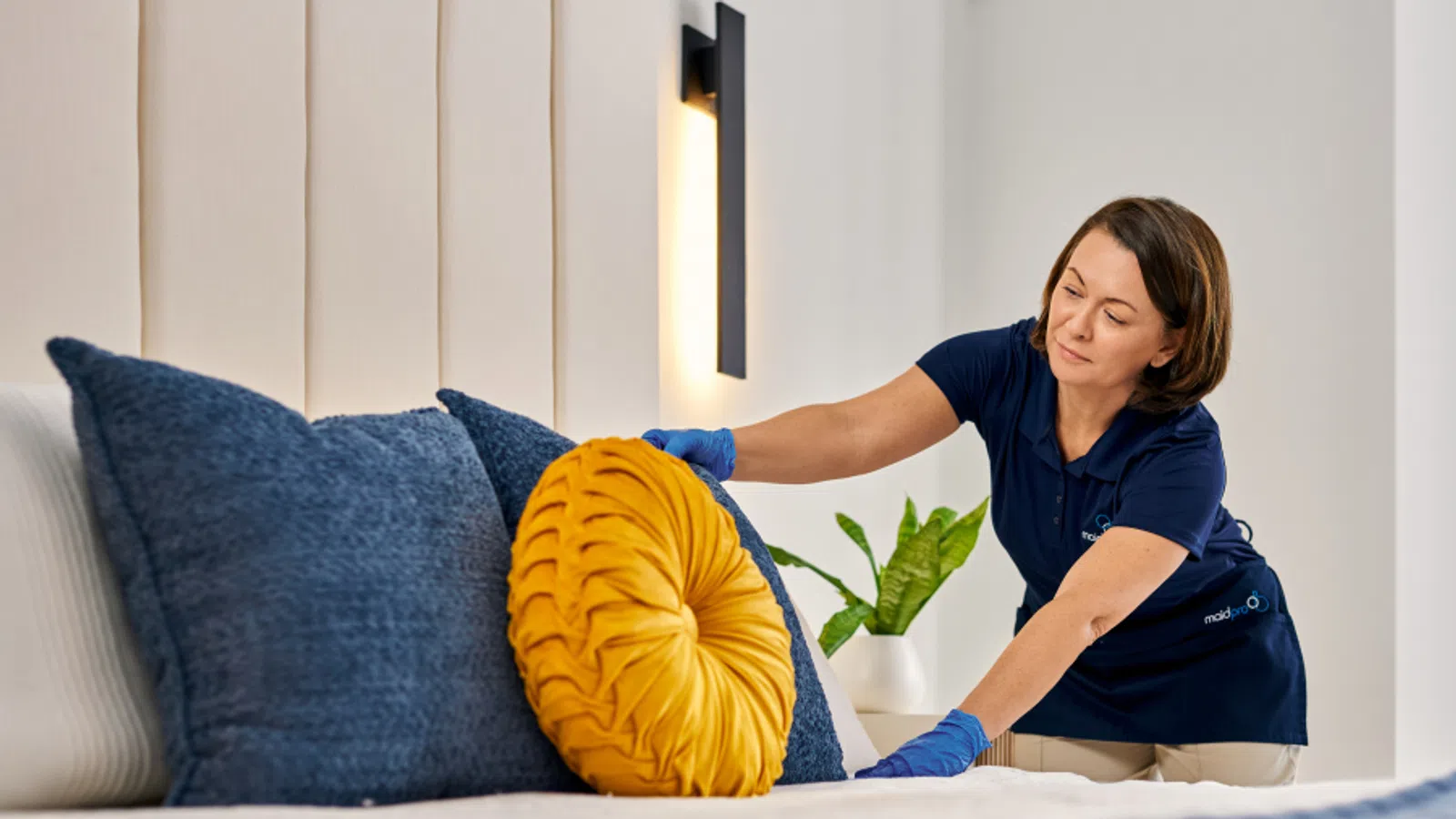 Professional cleaner arranging mustard yellow and navy blue pillows on a bed in a modern bedroom.