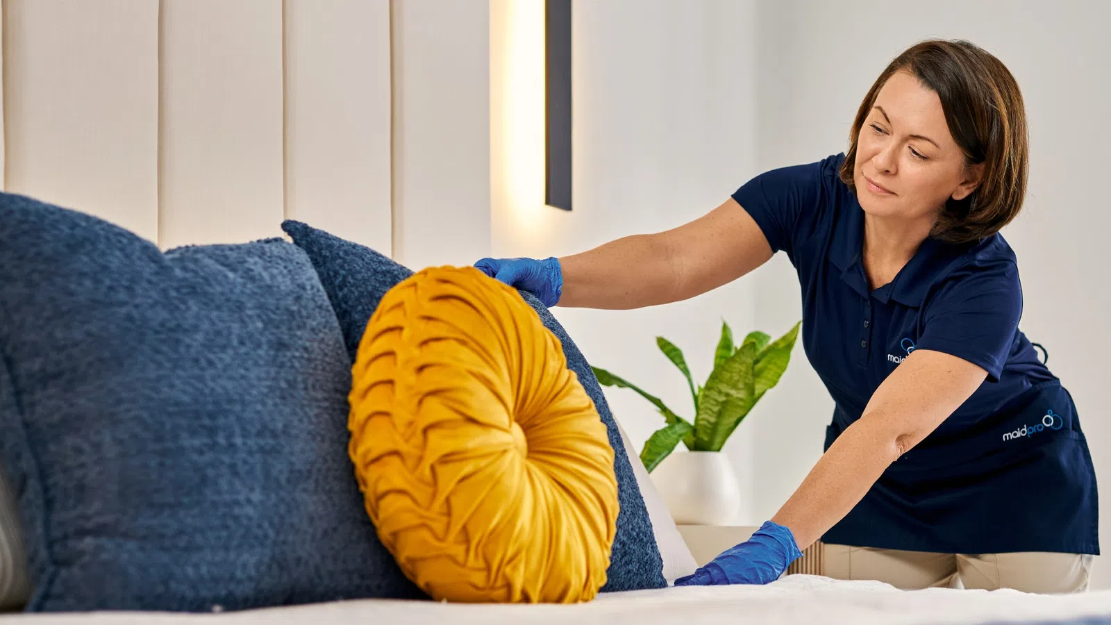 Housekeeper arranging bright yellow pillow on blue cushions on a neatly made bed in a modern room.
