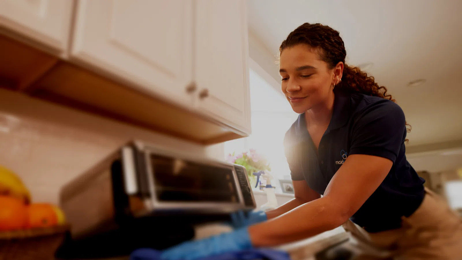Professional cleaner wiping kitchen countertop near toaster oven with blue cloth and gloves
