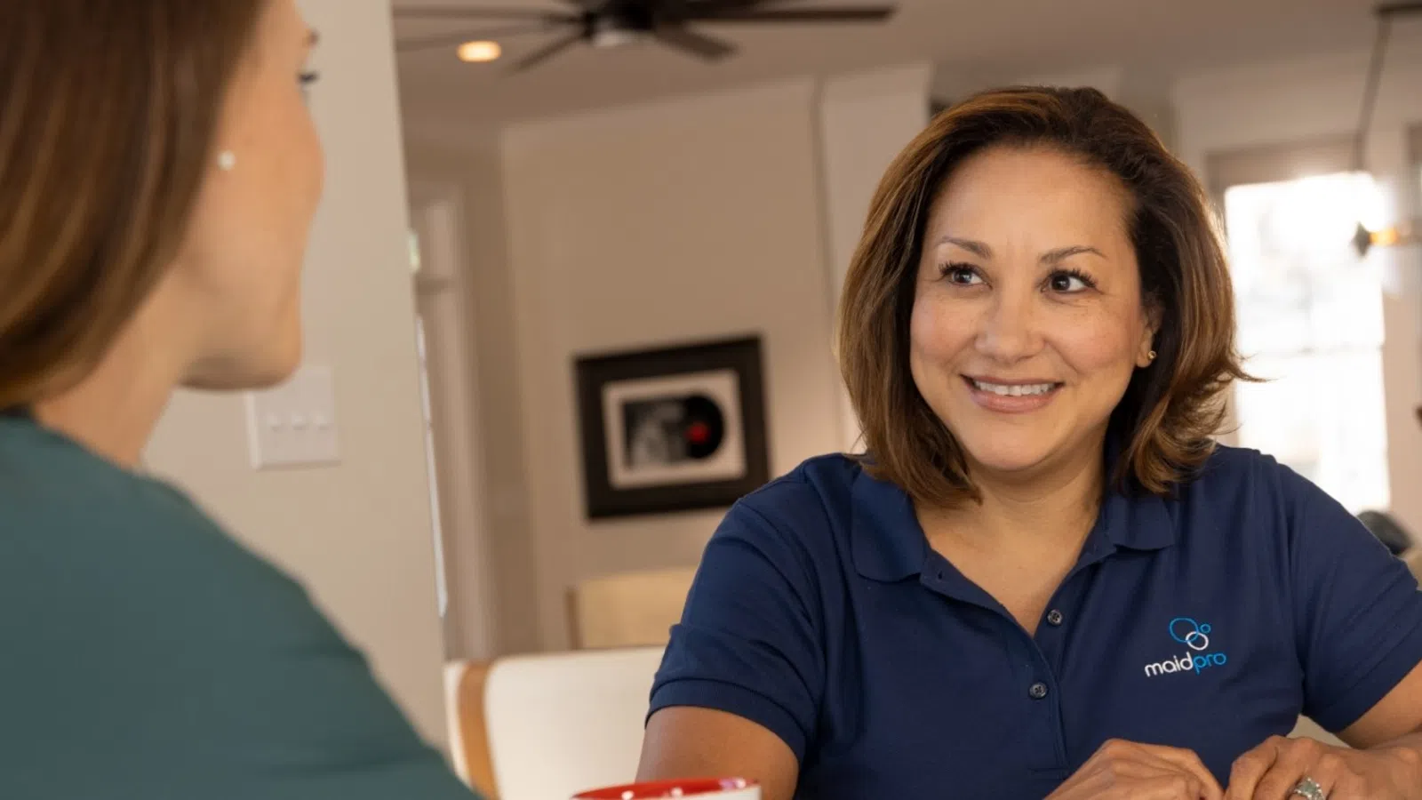 Smiling woman in a MaidPro polo shirt chatting with another woman at a table in a cozy home setting