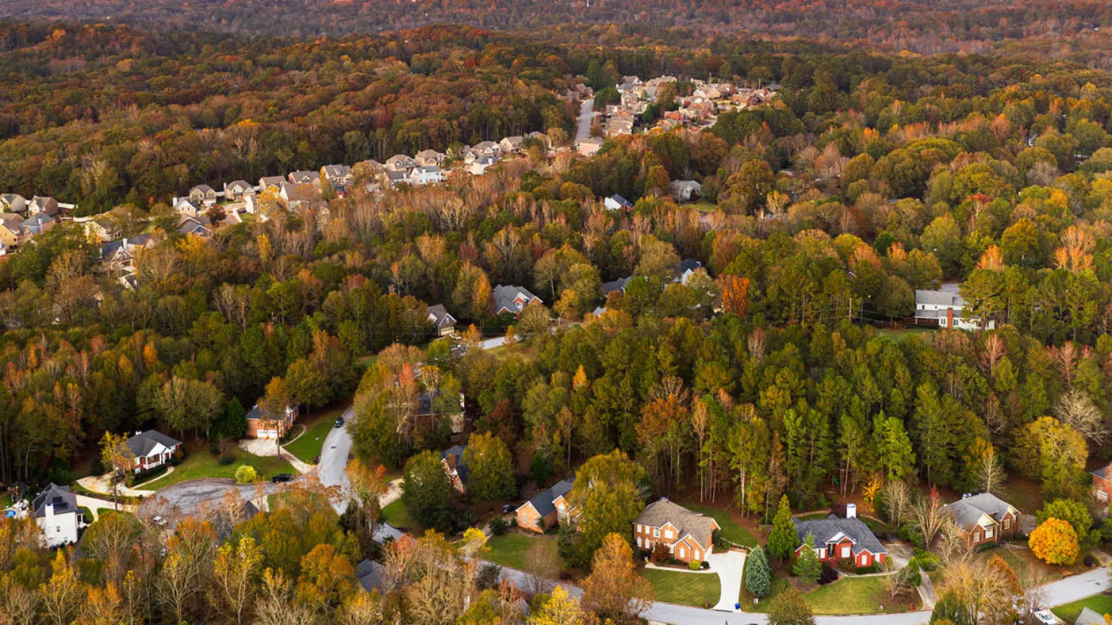 Aerial view of a suburban neighborhood surrounded by dense forest with autumn foliage under a clear sky