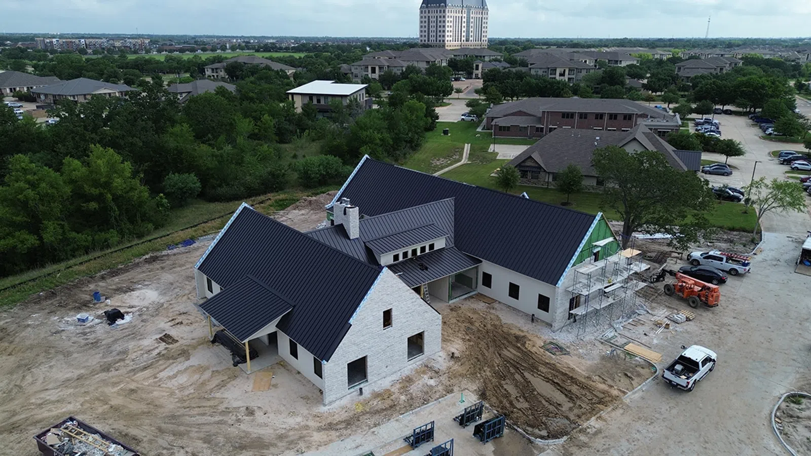 Aerial view of a modern house under construction with dark roofs surrounded by dirt and nearby residential buildings.