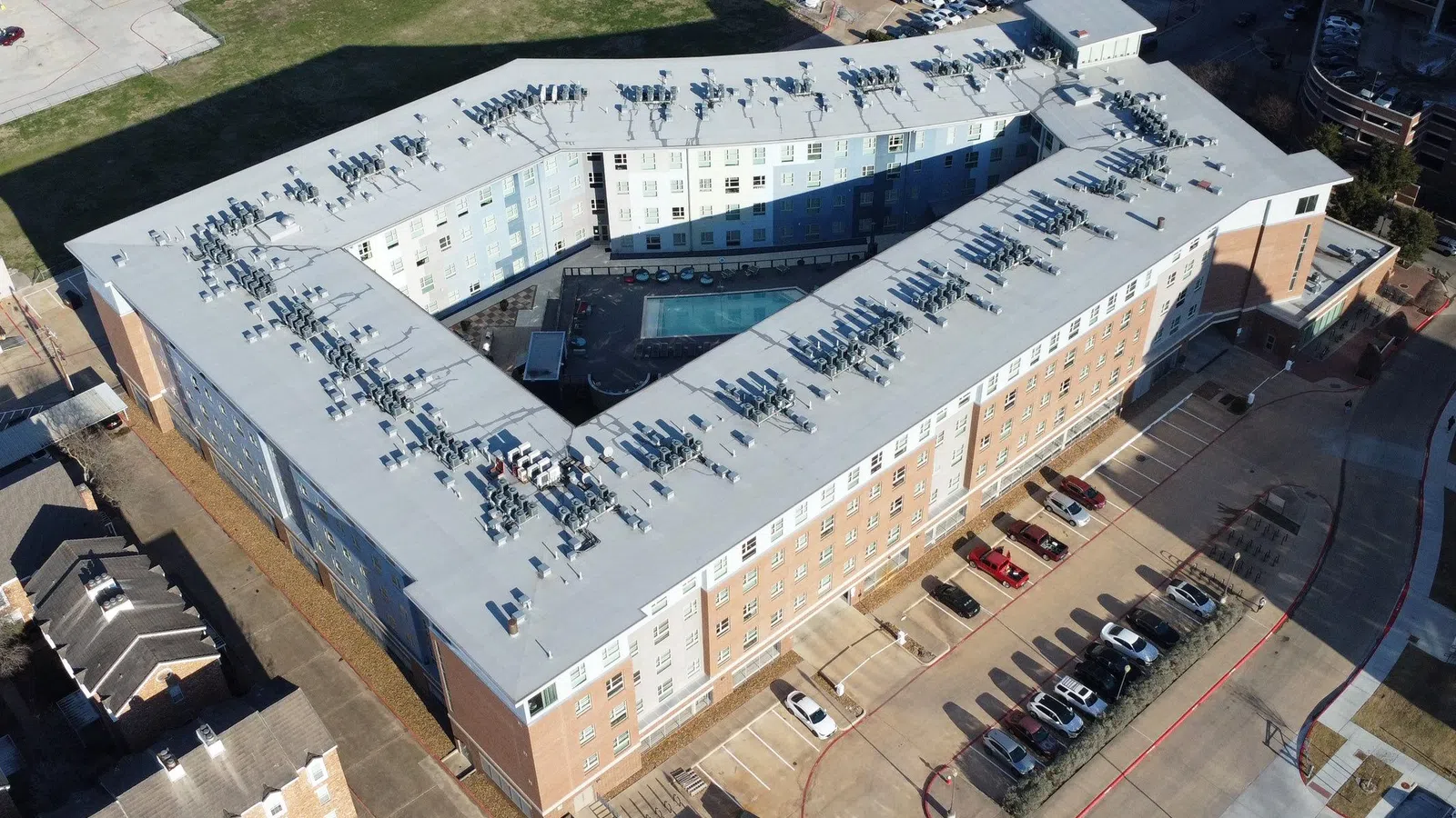 Aerial view of a large U-shaped apartment building with a central pool and surrounding parking lot on a sunny day