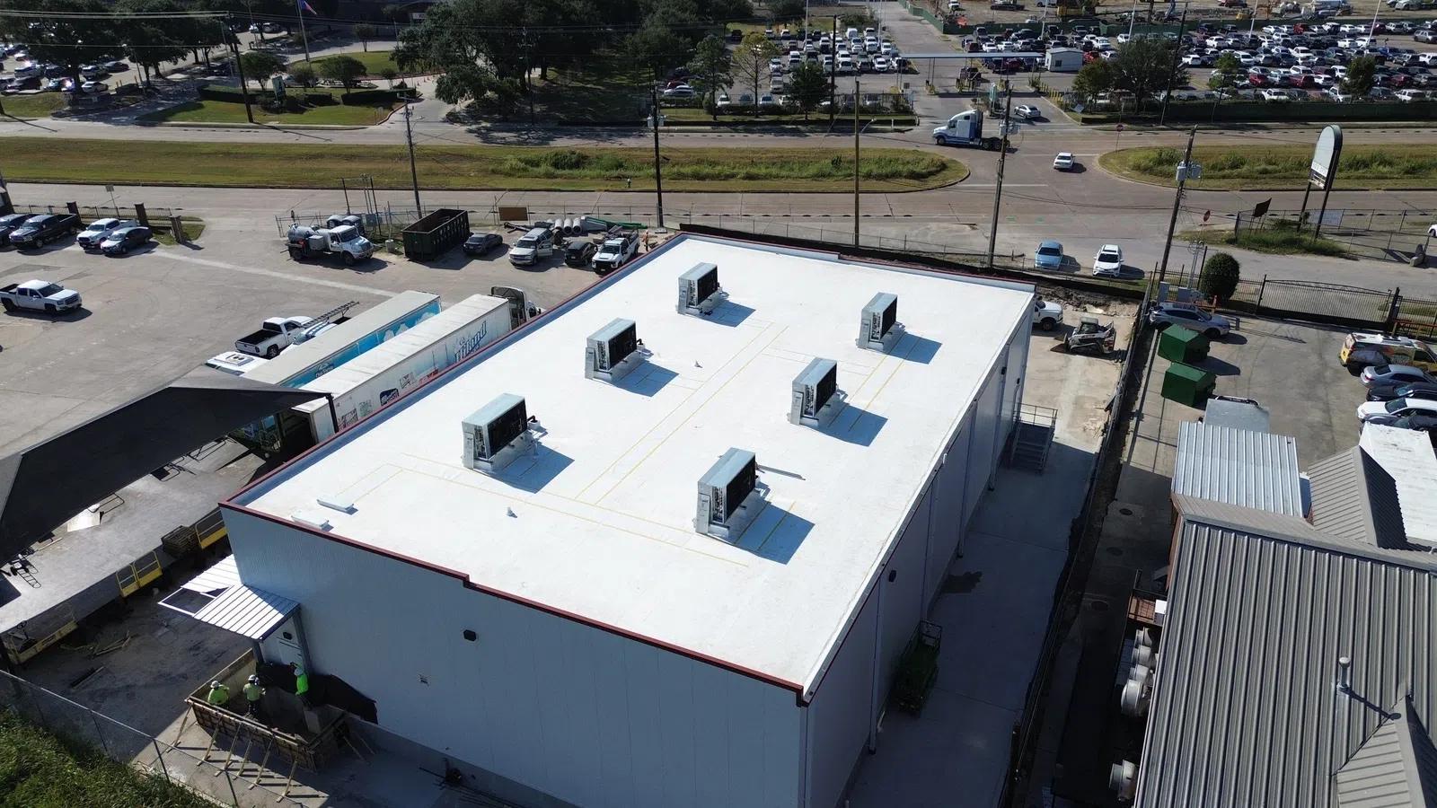 Aerial view of a completed white TPO membrane roof on a commercial industrial building in the Houston area, with six rooftop HVAC units, a loading dock, and a Mattco crew working on site surrounded by truck and trailer traffic.