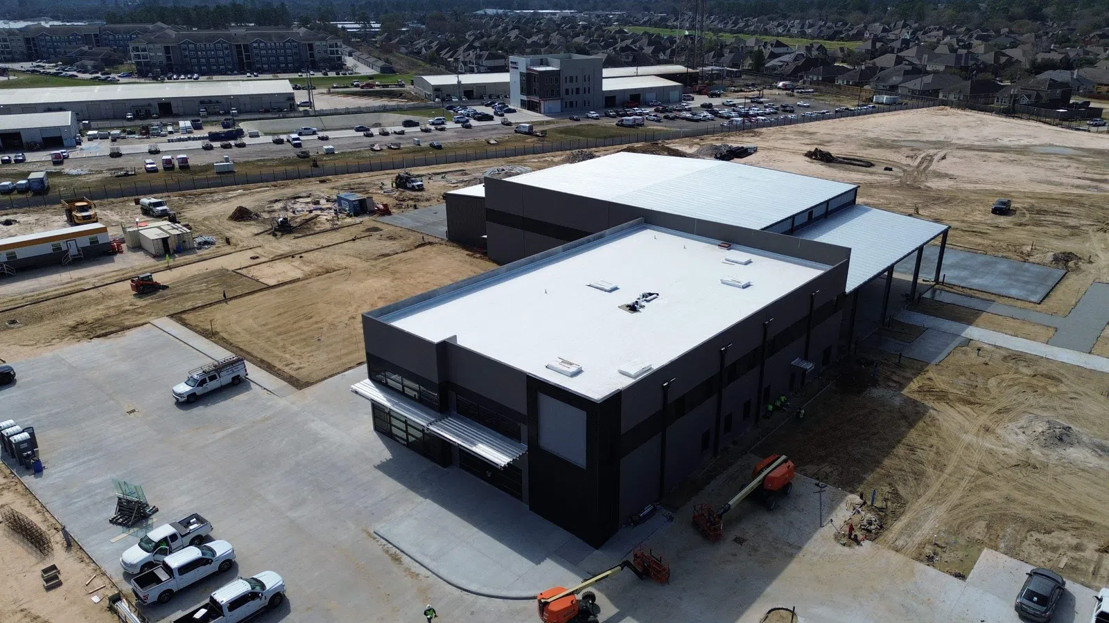 Aerial view of a large commercial and industrial facility under construction in the Houston area, showing a combination of white TPO membrane roofing and standing seam metal roofing across multiple connected building sections with surrounding development.