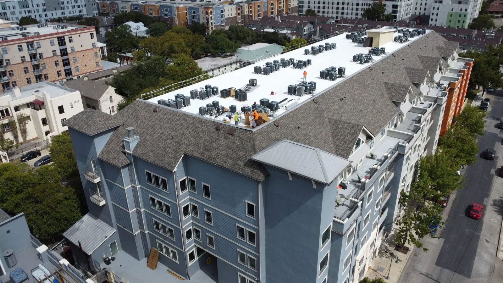 Aerial view of a mixed roof system on a multi-family student housing building in Austin, Texas, showing a white flat membrane roof section surrounded by architectural shingle roofing, with rows of rooftop HVAC units and a Mattco crew working on site near the University of Texas.