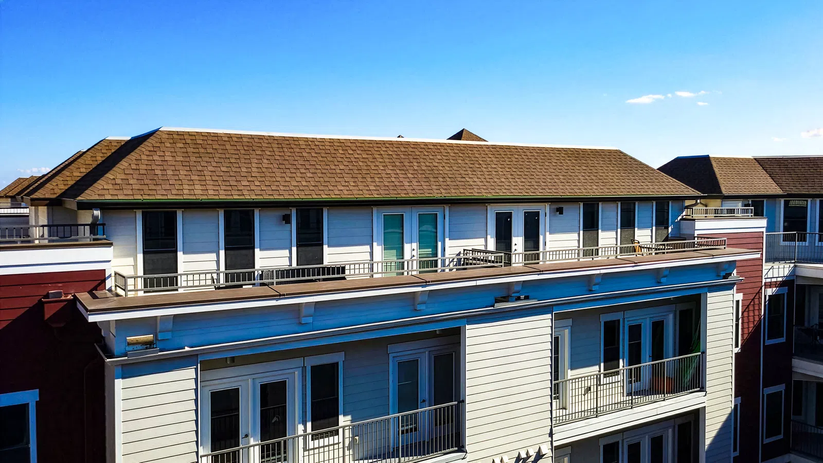 Close-up aerial view of a multifamily residential building with a shingle roof and rooftop balcony units against a clear blue sky.
