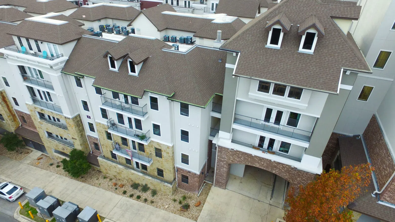 Aerial view of a large multifamily housing complex with shingle roofs and rooftop HVAC units, surrounded by fall foliage and city buildings.
