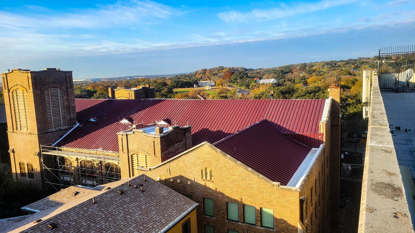 Aerial view of a historic brick church building with a dark red standing seam metal roof, with scaffolding on an adjacent structure and rolling tree-covered hills in the background.