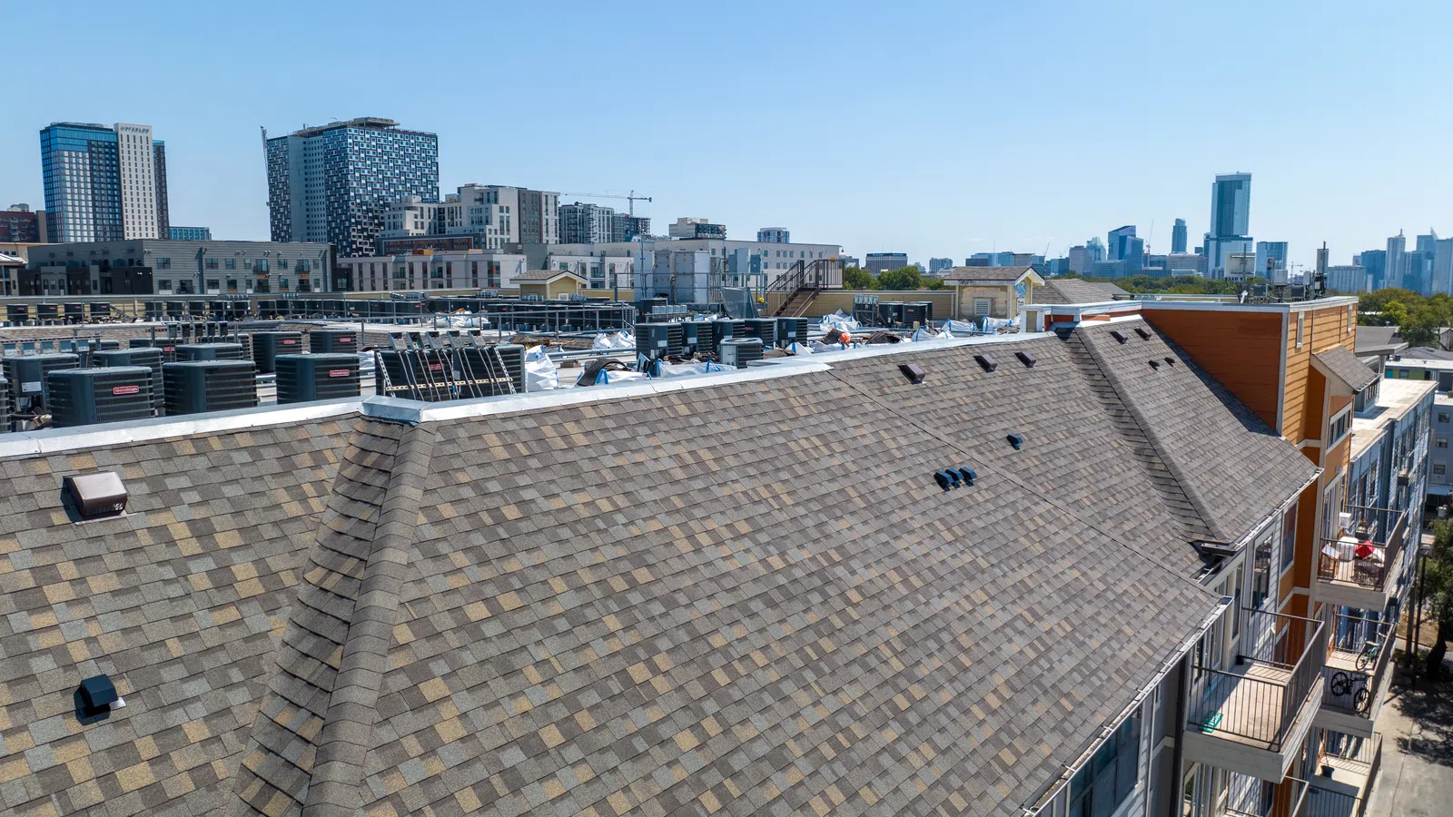 Aerial view of a completed architectural shingle roof on a multi-family student housing property in Austin, Texas, with the downtown Austin skyline visible in the background and rooftop HVAC equipment on an adjacent building.