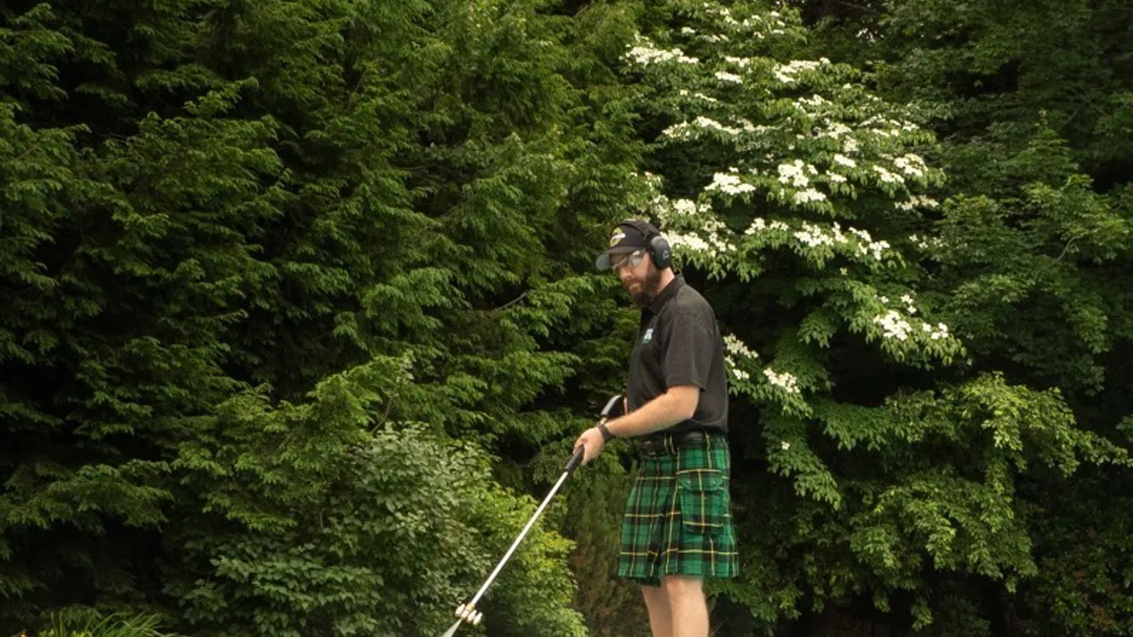 Man wearing a kilt pressure washing the edge of a swimming pool surrounded by greenery and trees