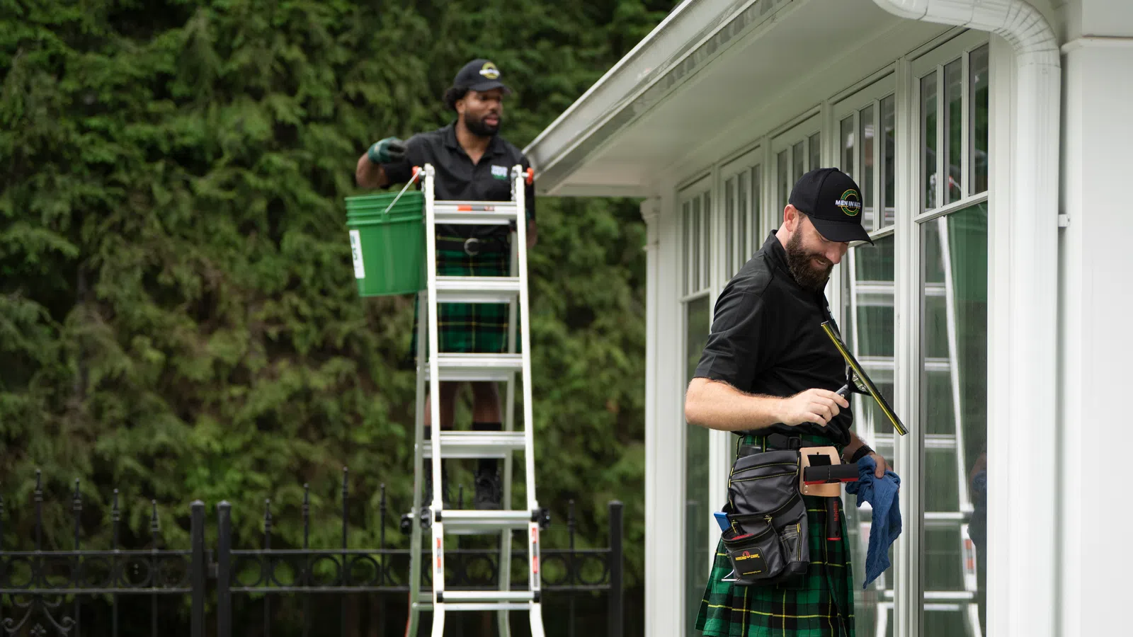 Two workers wearing kilts cleaning large windows outside a house, one on ladder, one wiping glass with squeegee.