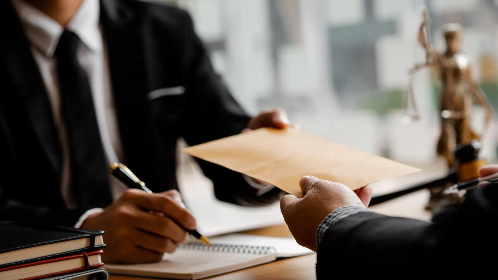 Two professionals in suits exchanging a brown envelope during a legal or business meeting with documents and scales of justice.
