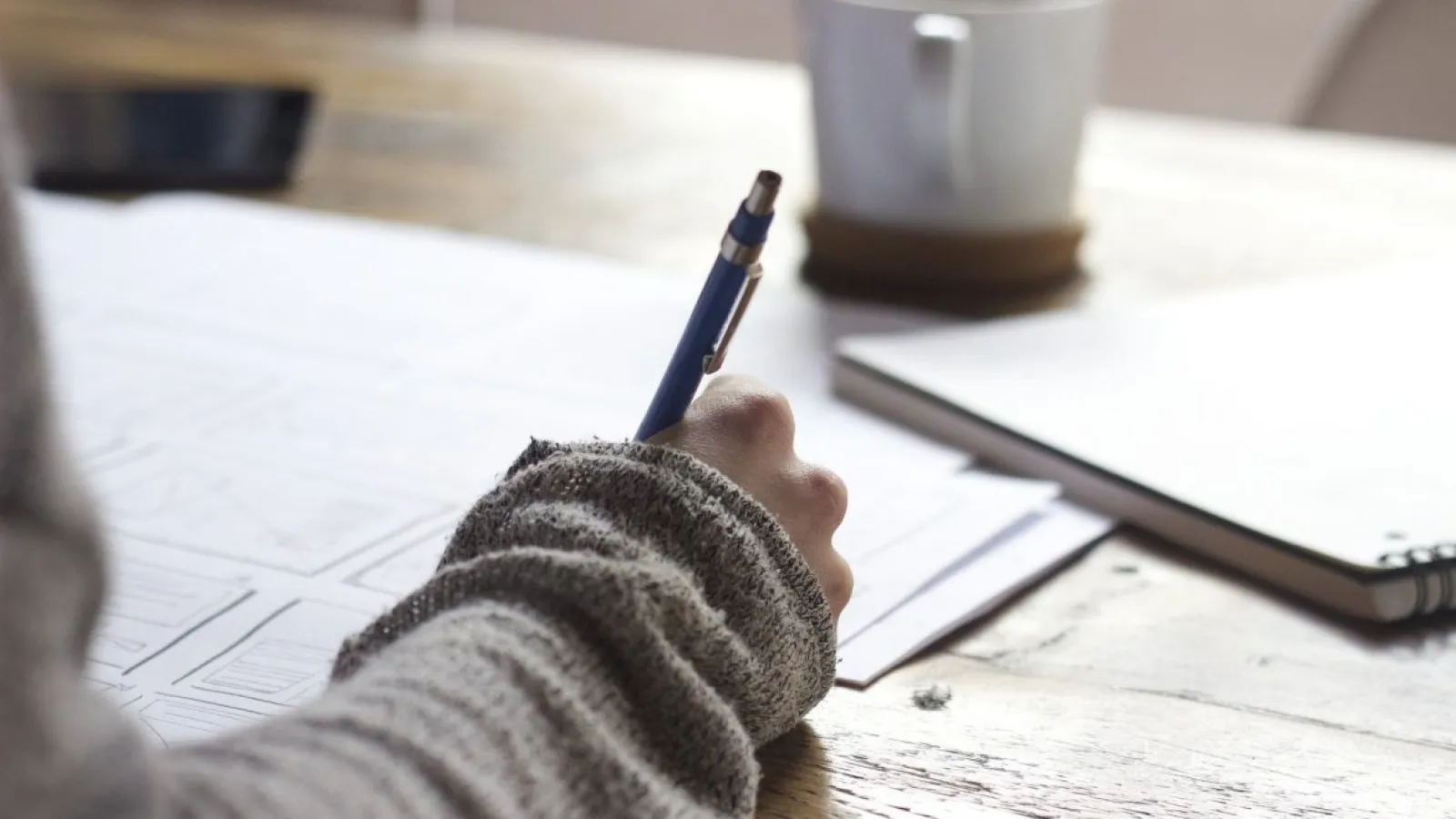 Hand writing with blue pen on paper at wooden table with notebook and coffee cup in soft background