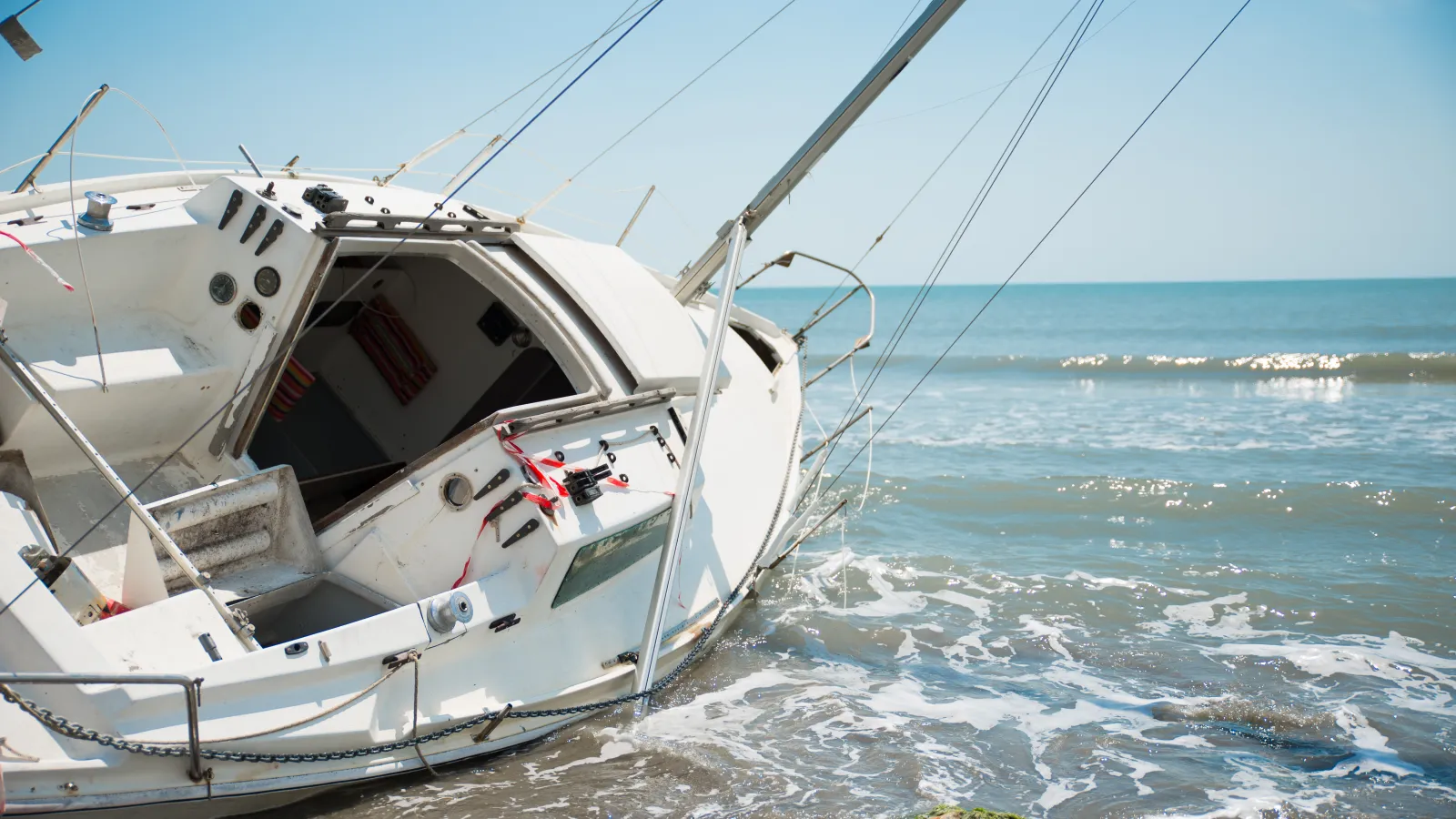 White sailboat partially submerged and tilted on rocky shoreline under clear blue sky on calm sea