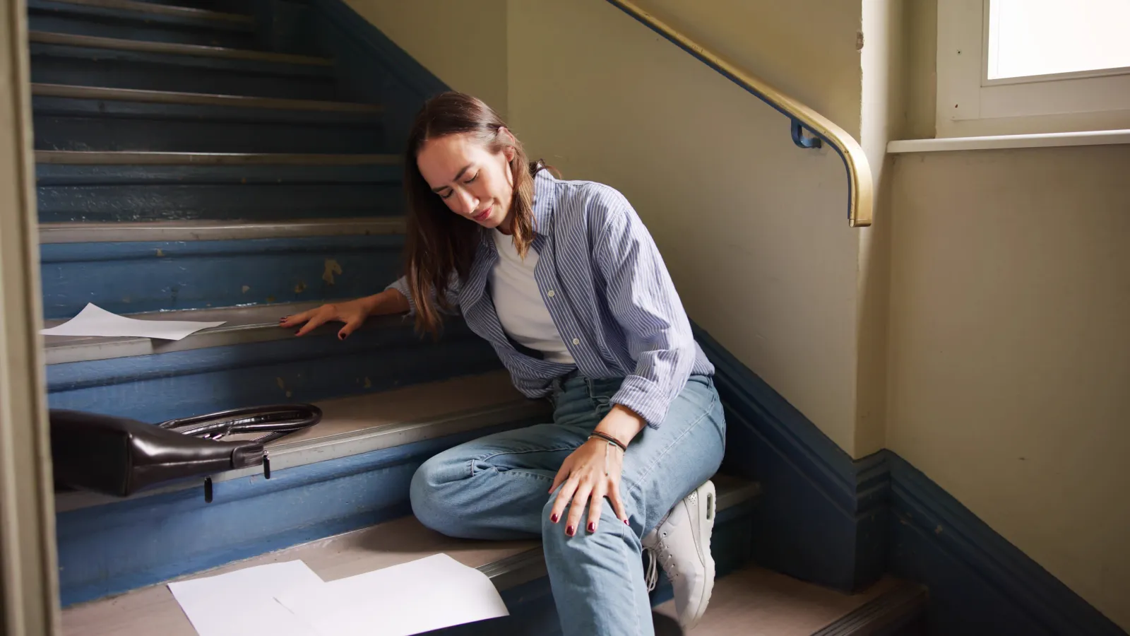Young woman sitting on stairs holding her knee in pain surrounded by scattered papers and a briefcase.