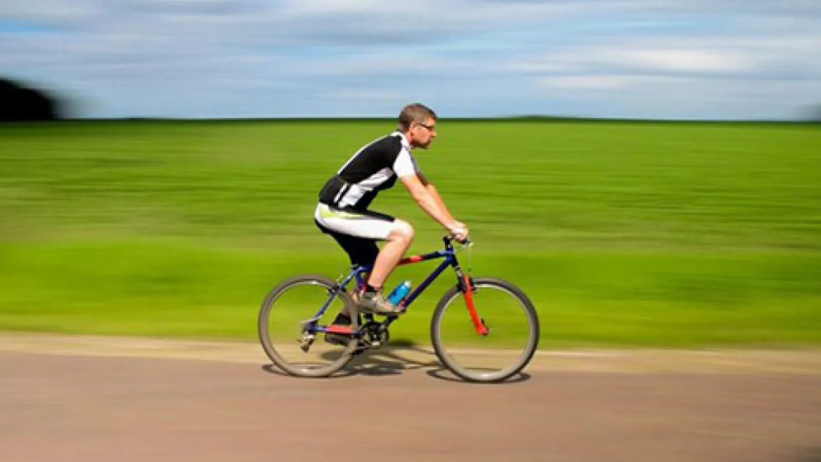Cyclist in black and white gear rides a bike fast along a rural road with green fields in the background