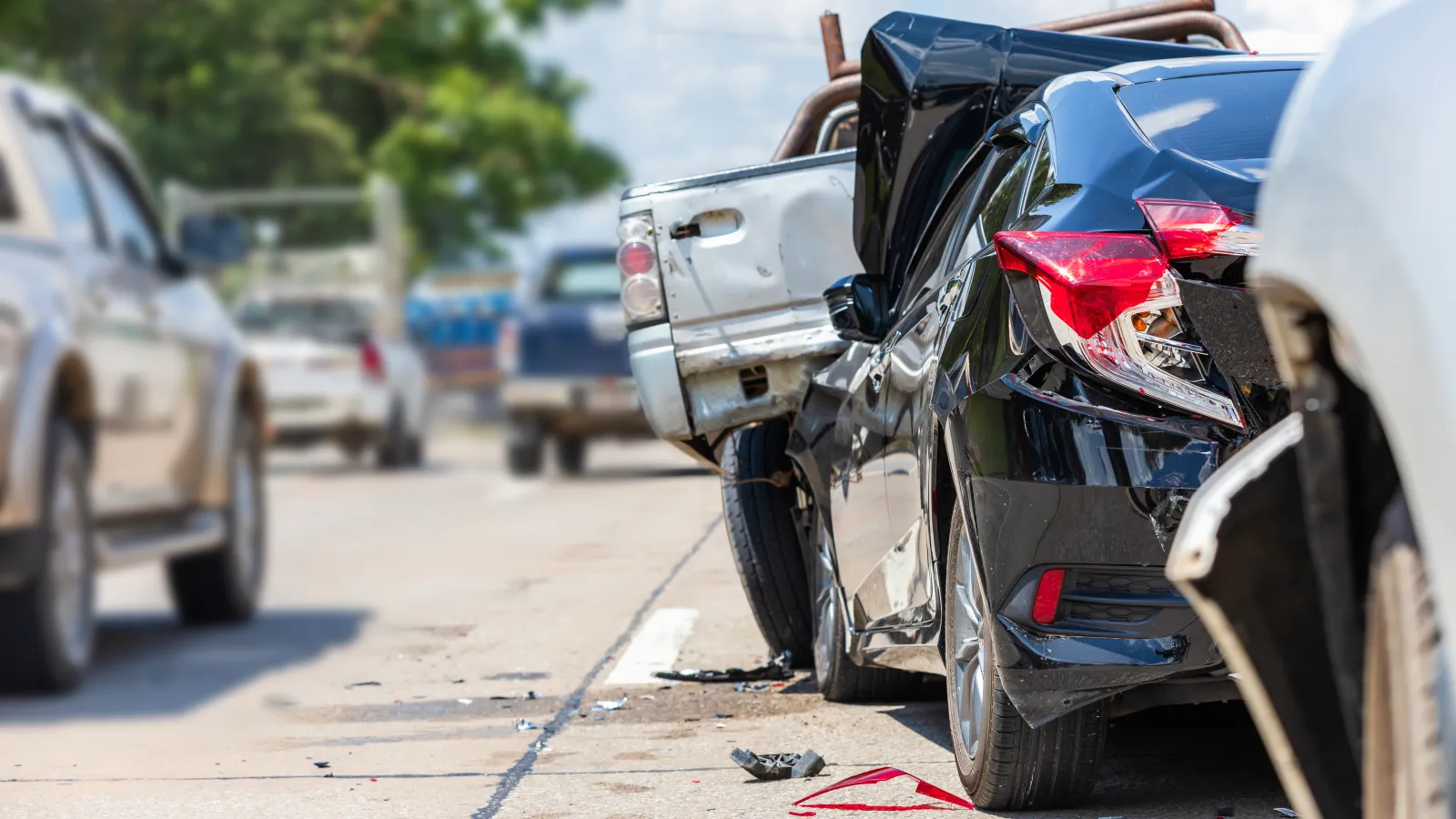 Damaged black car and white pickup truck involved in a rear-end collision on a street with traffic.