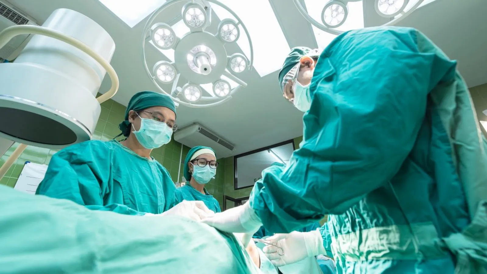 Surgical team in green scrubs performing an operation under bright operating room lights.