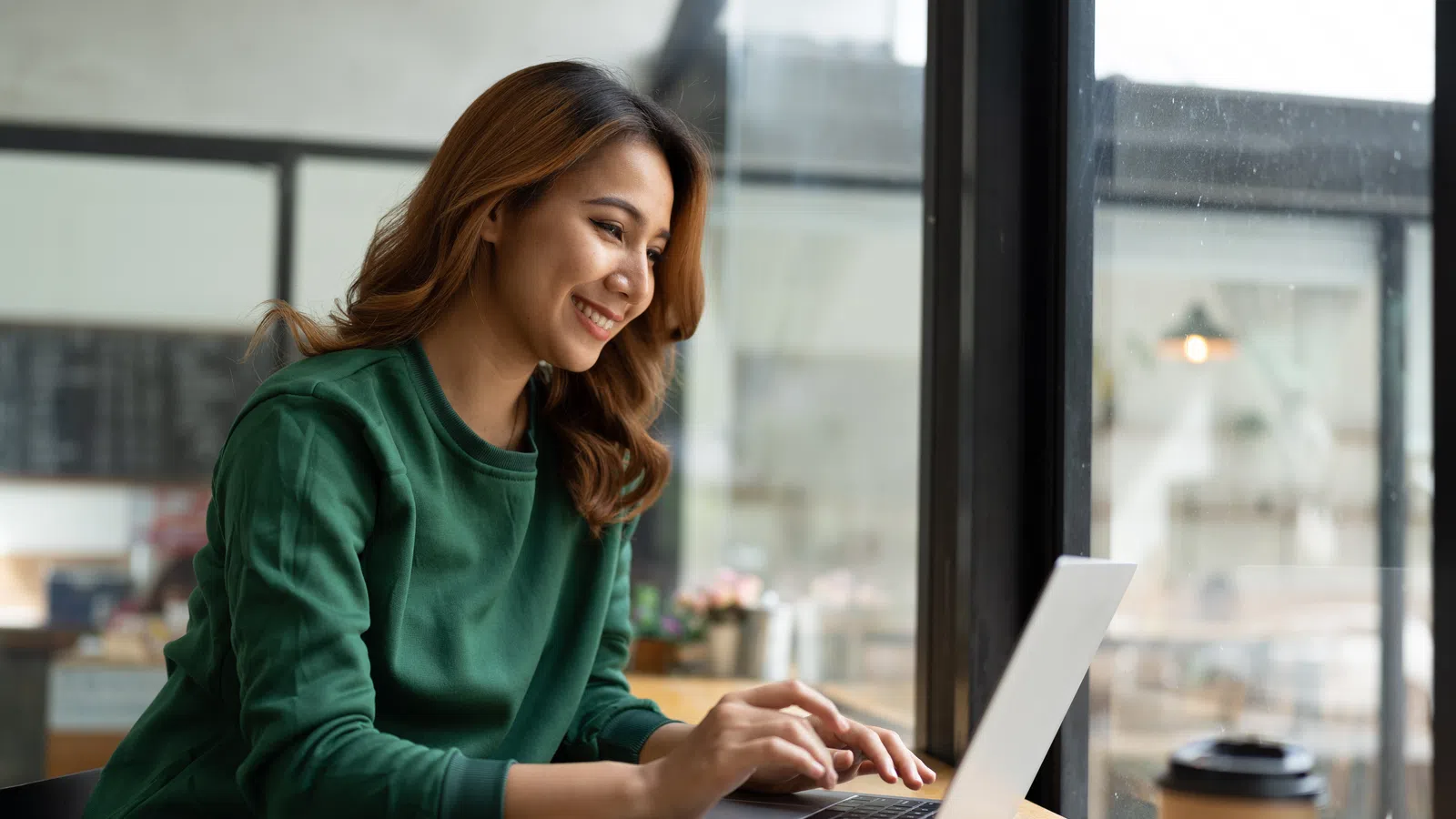 Young woman smiling working on laptop at cafe table near window with coffee cup and smartphone nearby