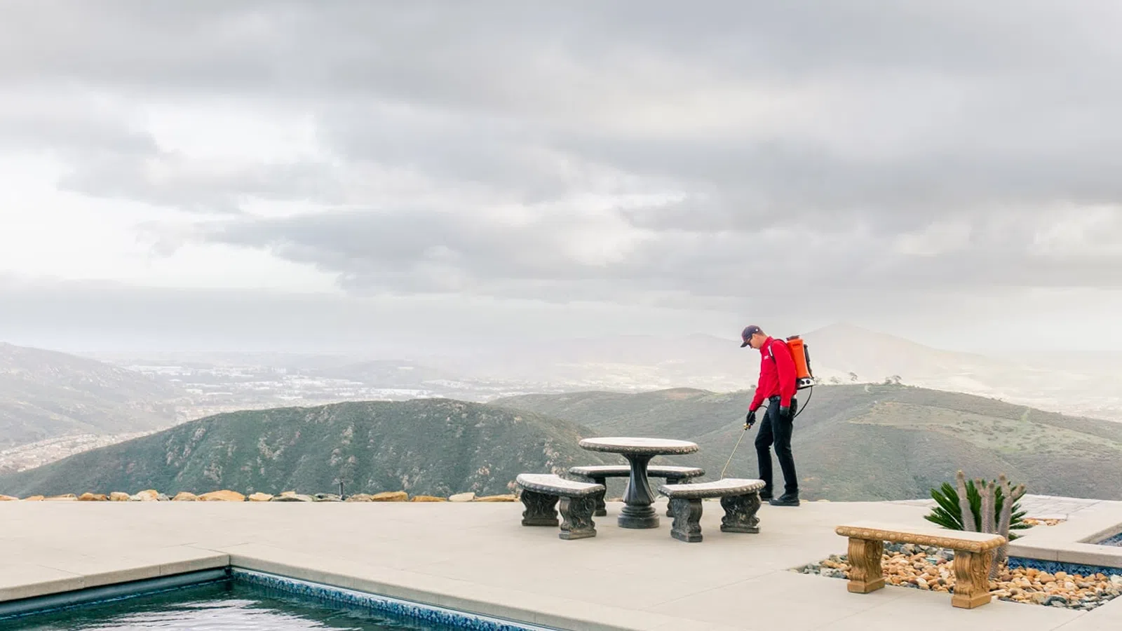Person in red jacket near outdoor stone table by pool with mountains under cloudy sky in background