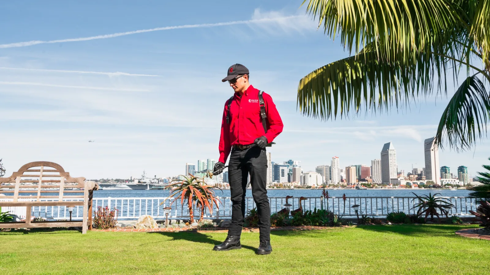Technician in red jacket inspecting lawn with city skyline and waterfront in background under clear sky