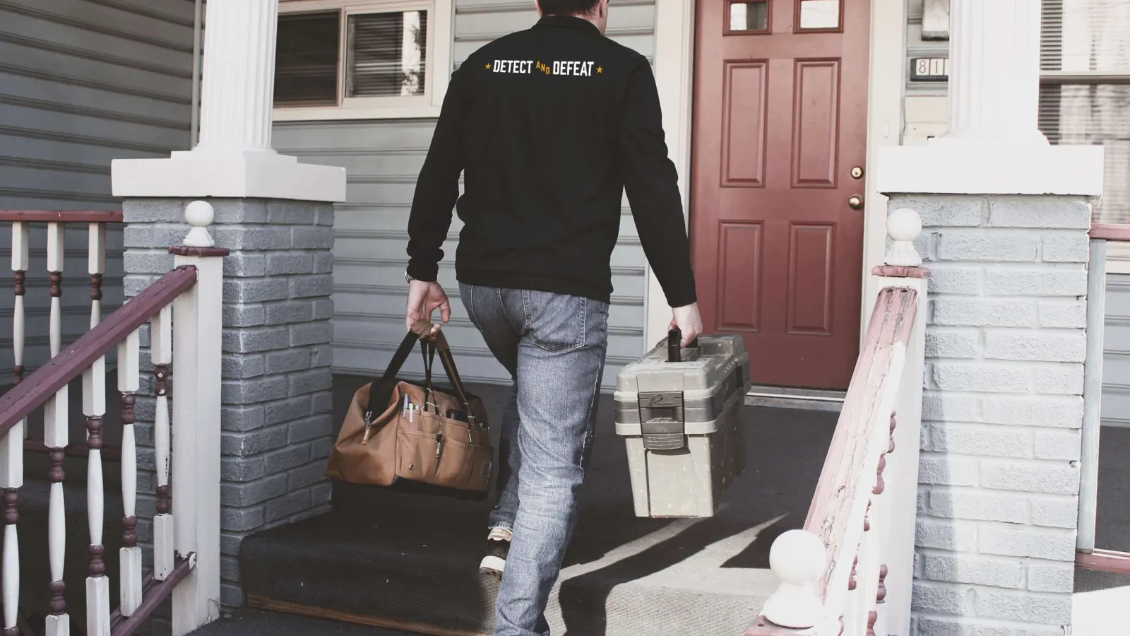 Man carrying tool and equipment bags walking up porch steps to house with red front door.