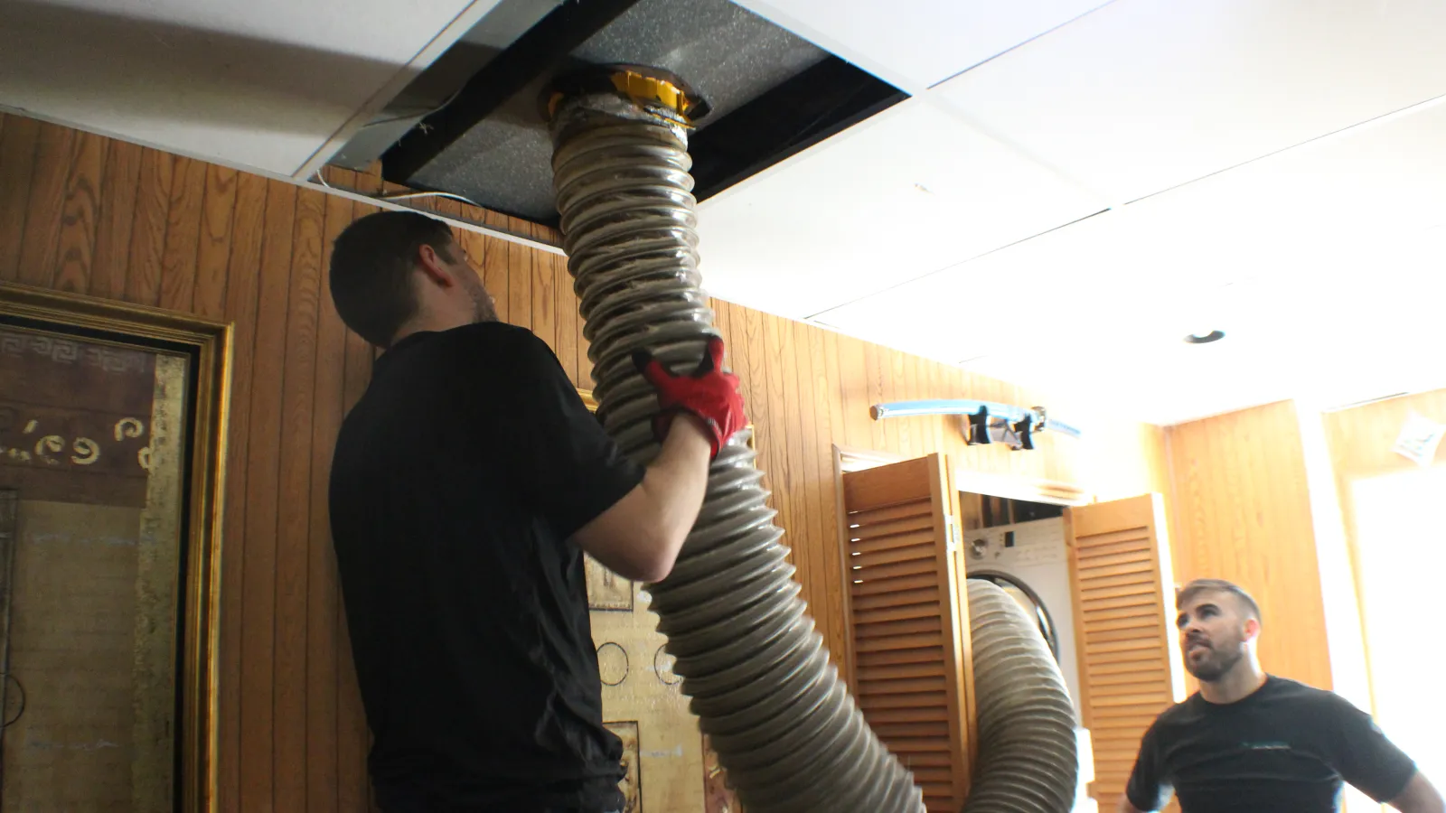 Two men installing a large flexible duct into a ceiling vent in a wood-paneled room.