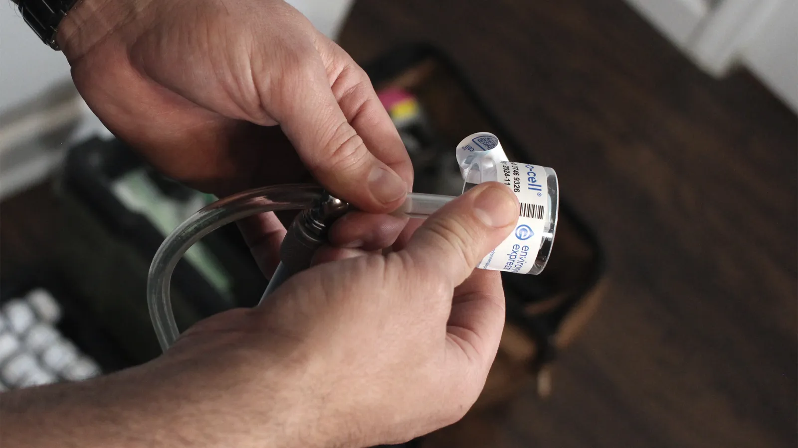 Close-up of hands holding and inspecting a gas detector sensor with tubing indoors on wooden floor