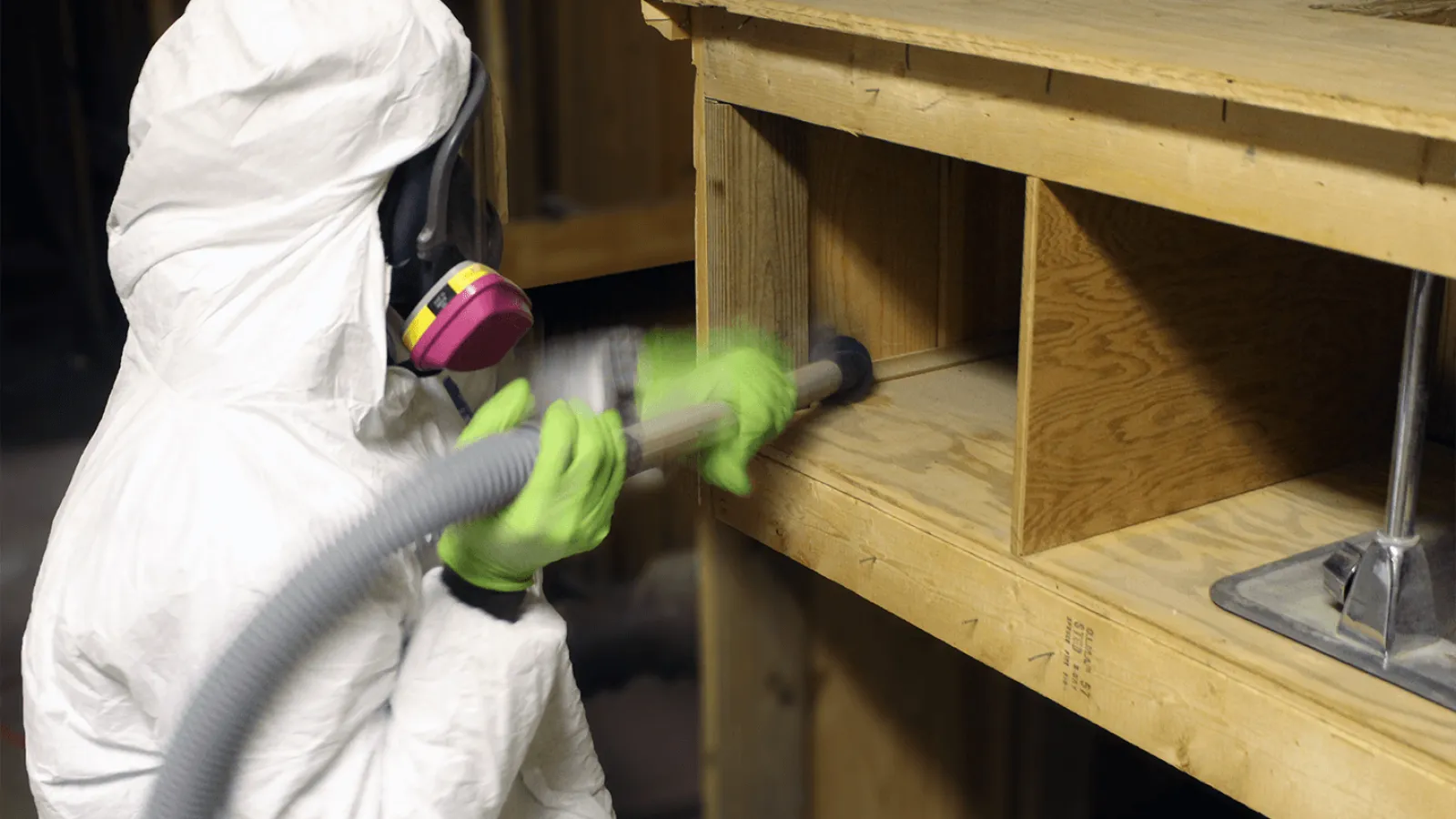 Person in protective gear using a vacuum hose to clean inside a wooden structure in a dimly lit area