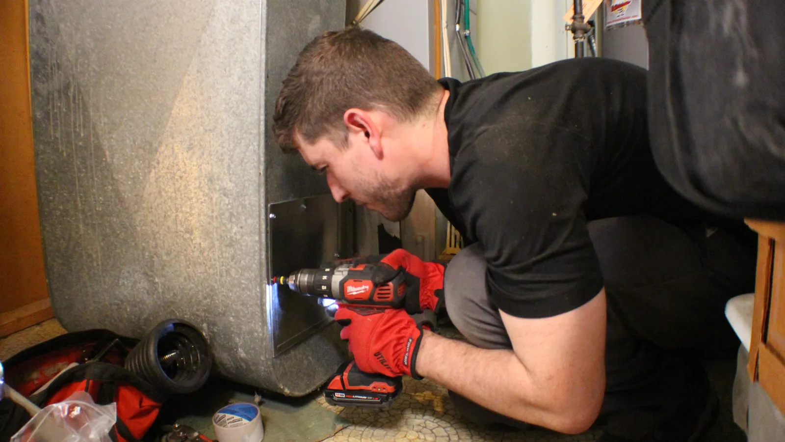 Technician in red gloves using a cordless drill to repair a large metal HVAC unit indoors.