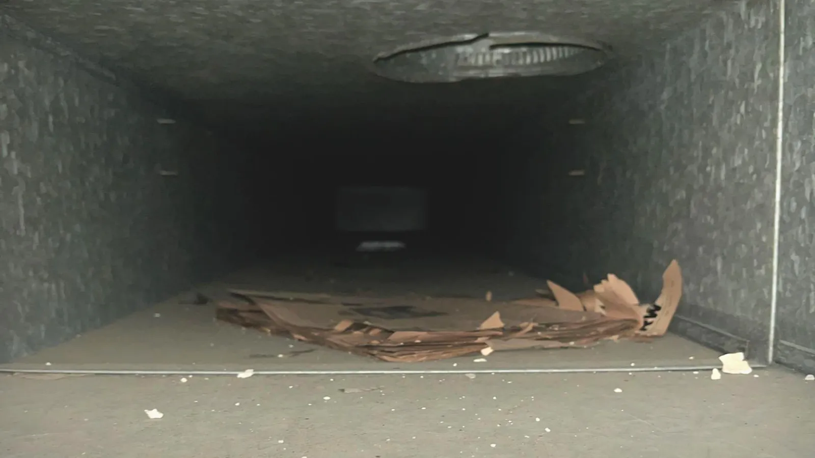 Inside view of a dusty, dirty HVAC duct with debris and rusted vent cover visible.