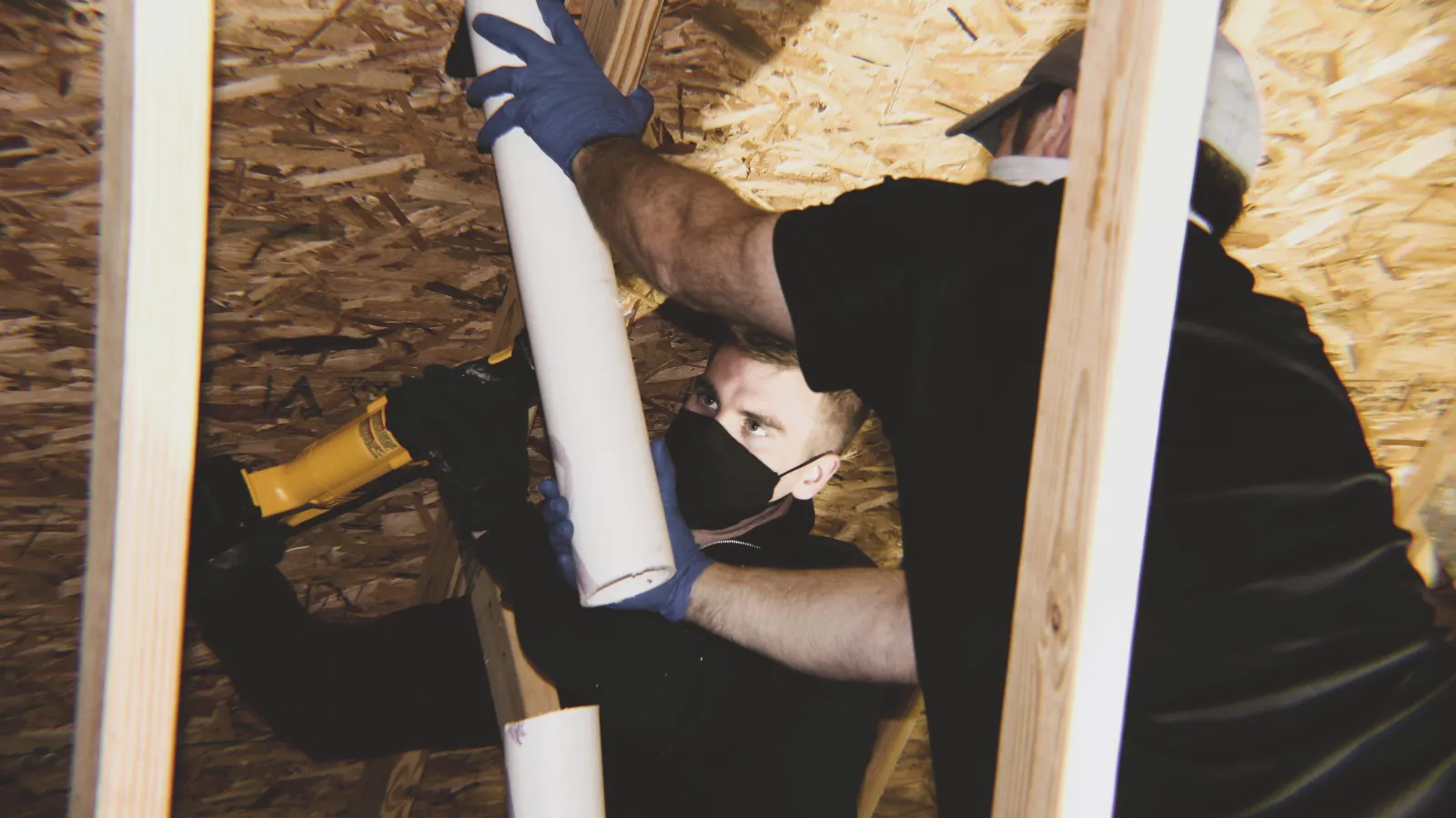 Two workers wearing masks and gloves installing PVC pipe in a wooden framed attic space.