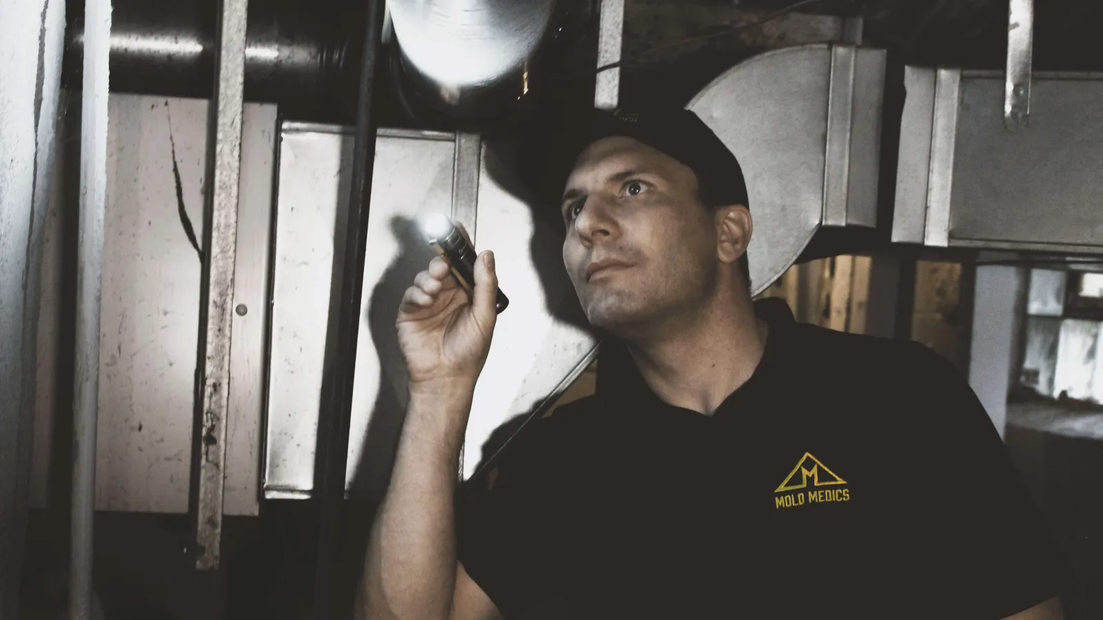 Technician inspecting HVAC ductwork indoors using a flashlight in low light conditions wearing a Mold Medics shirt
