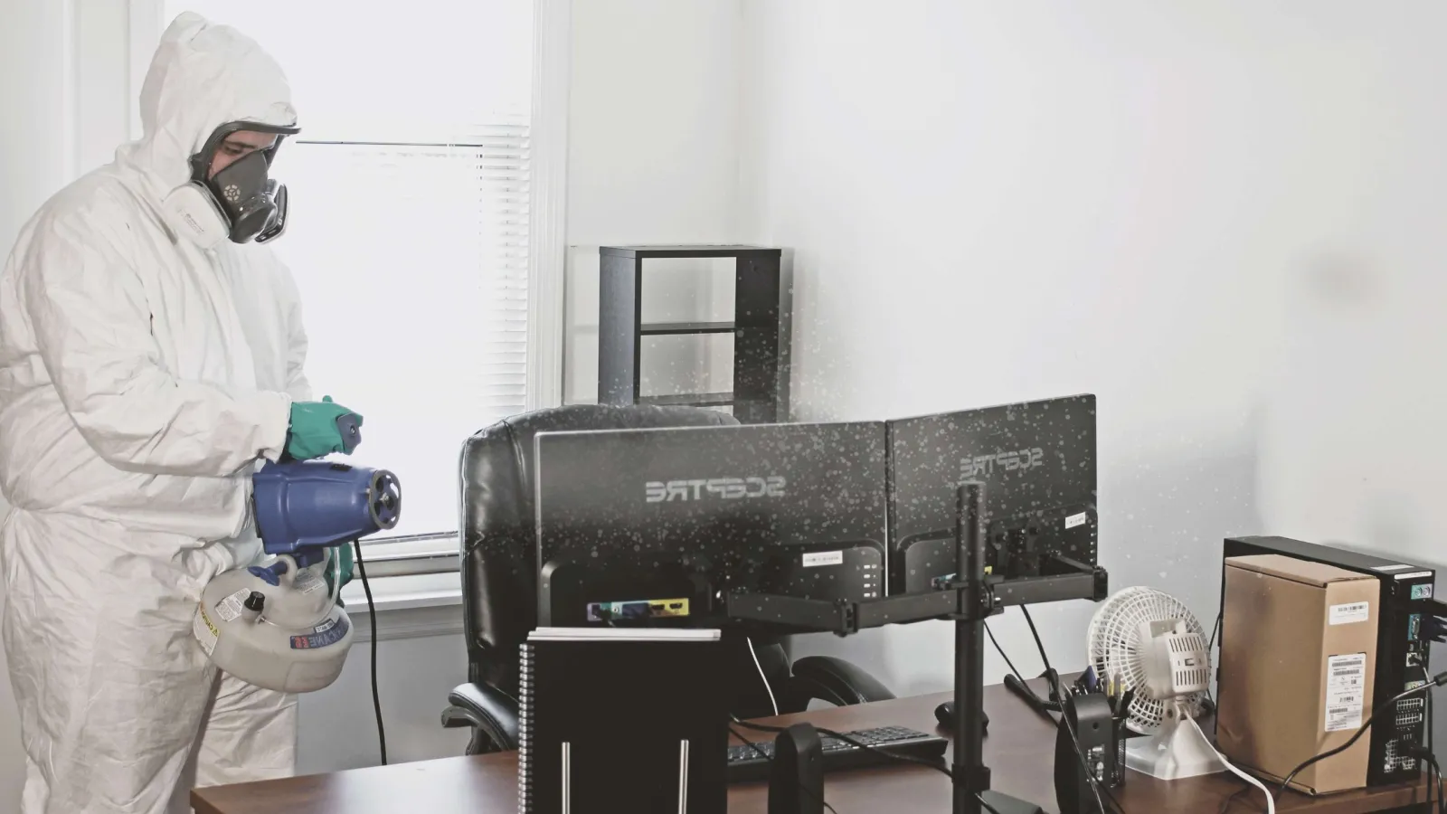 Person in full protective suit disinfecting an office desk and computer monitors in a clean workspace.
