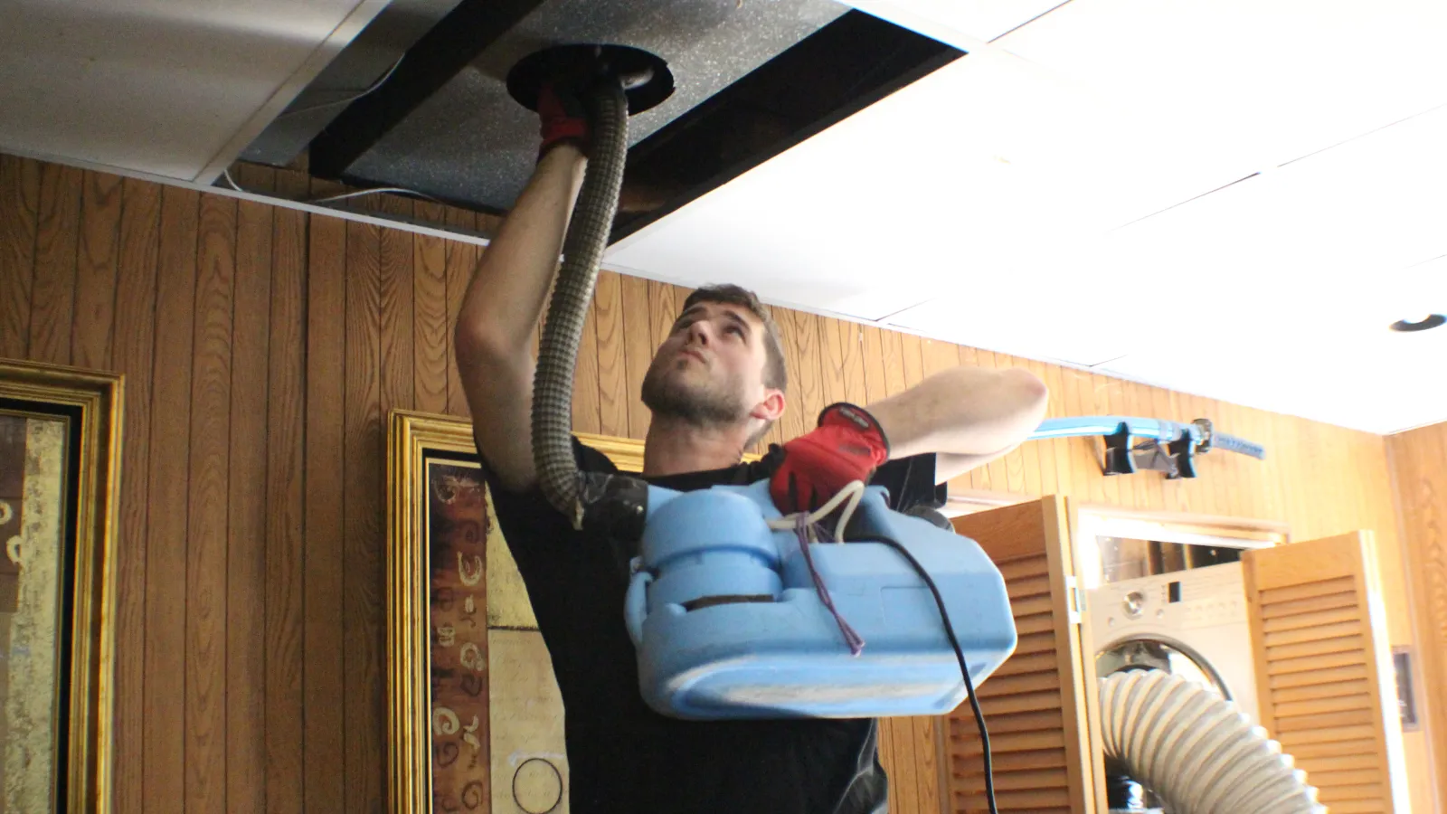 Technician using vacuum equipment to clean inside ceiling ducts in a residential room with wood panel walls.