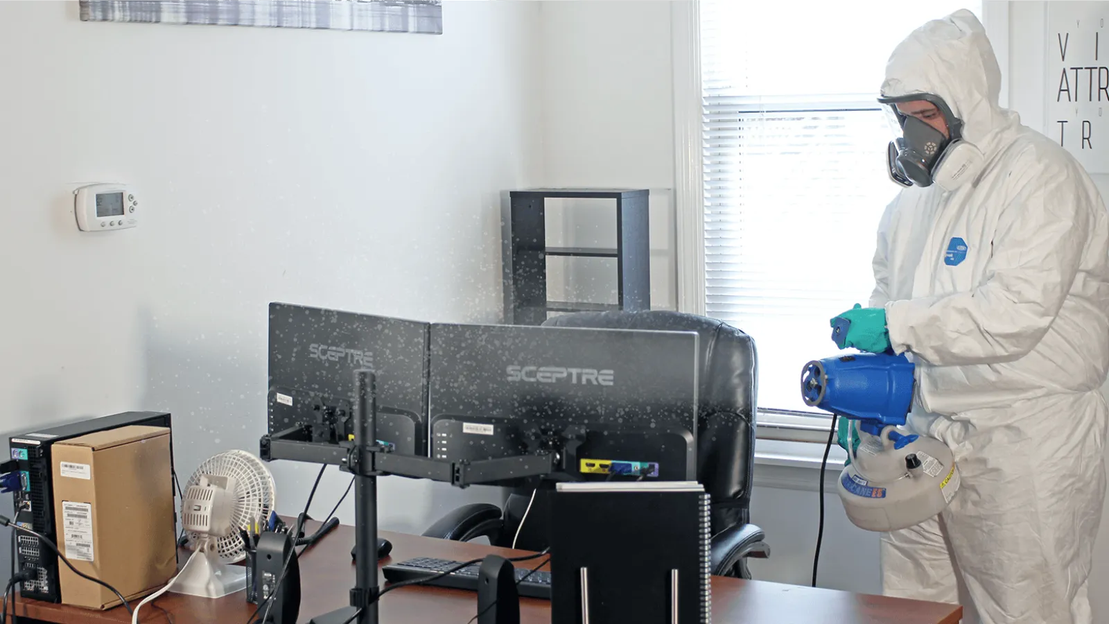 Person in protective suit disinfecting office space with computer monitors and desk equipment by window.