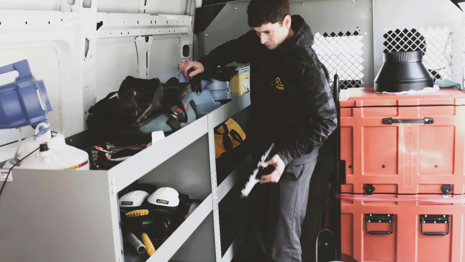 Worker organizing tools and equipment on shelves inside a white utility van with storage boxes and gear.
