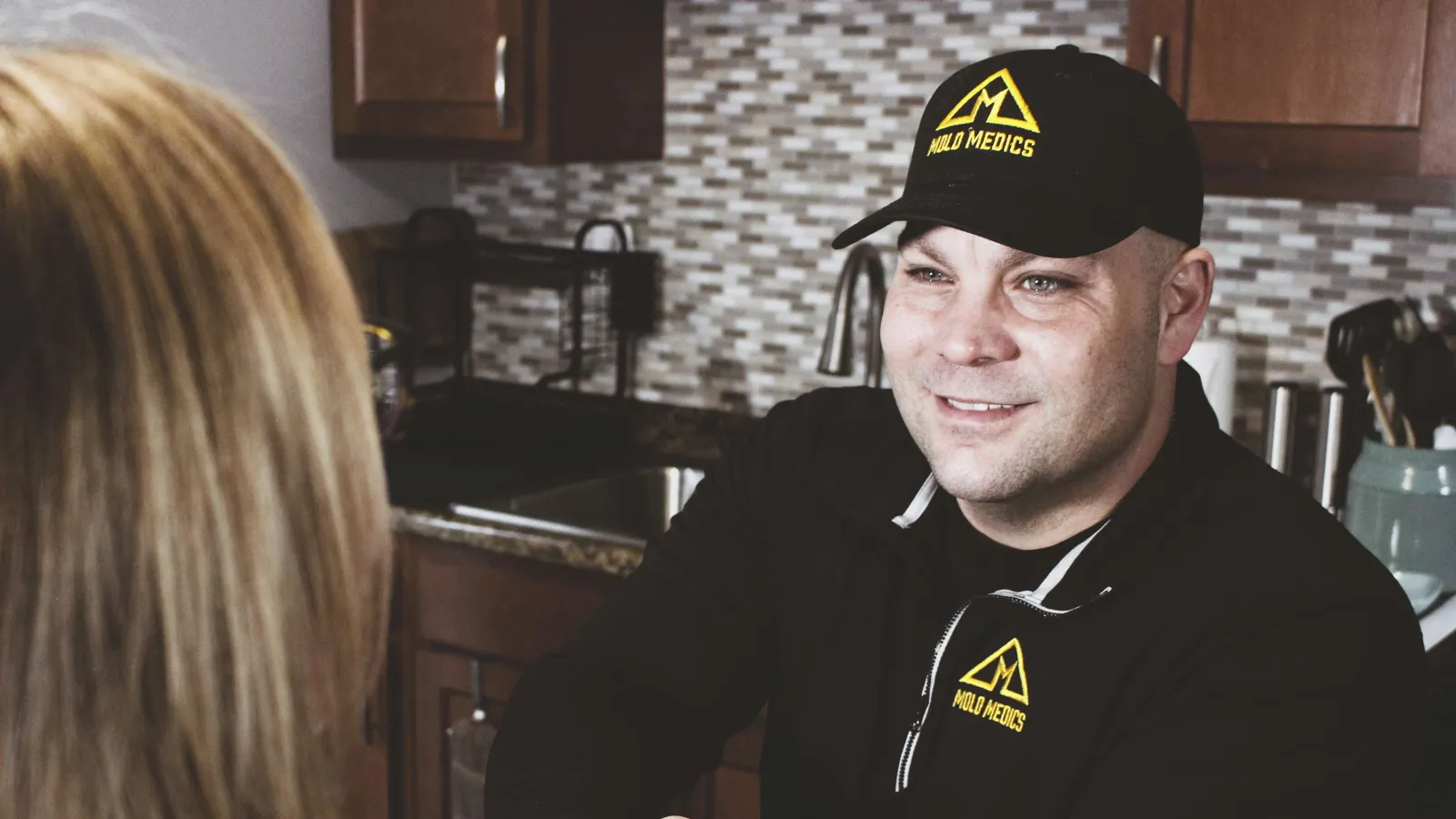 Mold Medics technician shaking hands with a customer in a kitchen setting, wearing branded black jacket and cap.