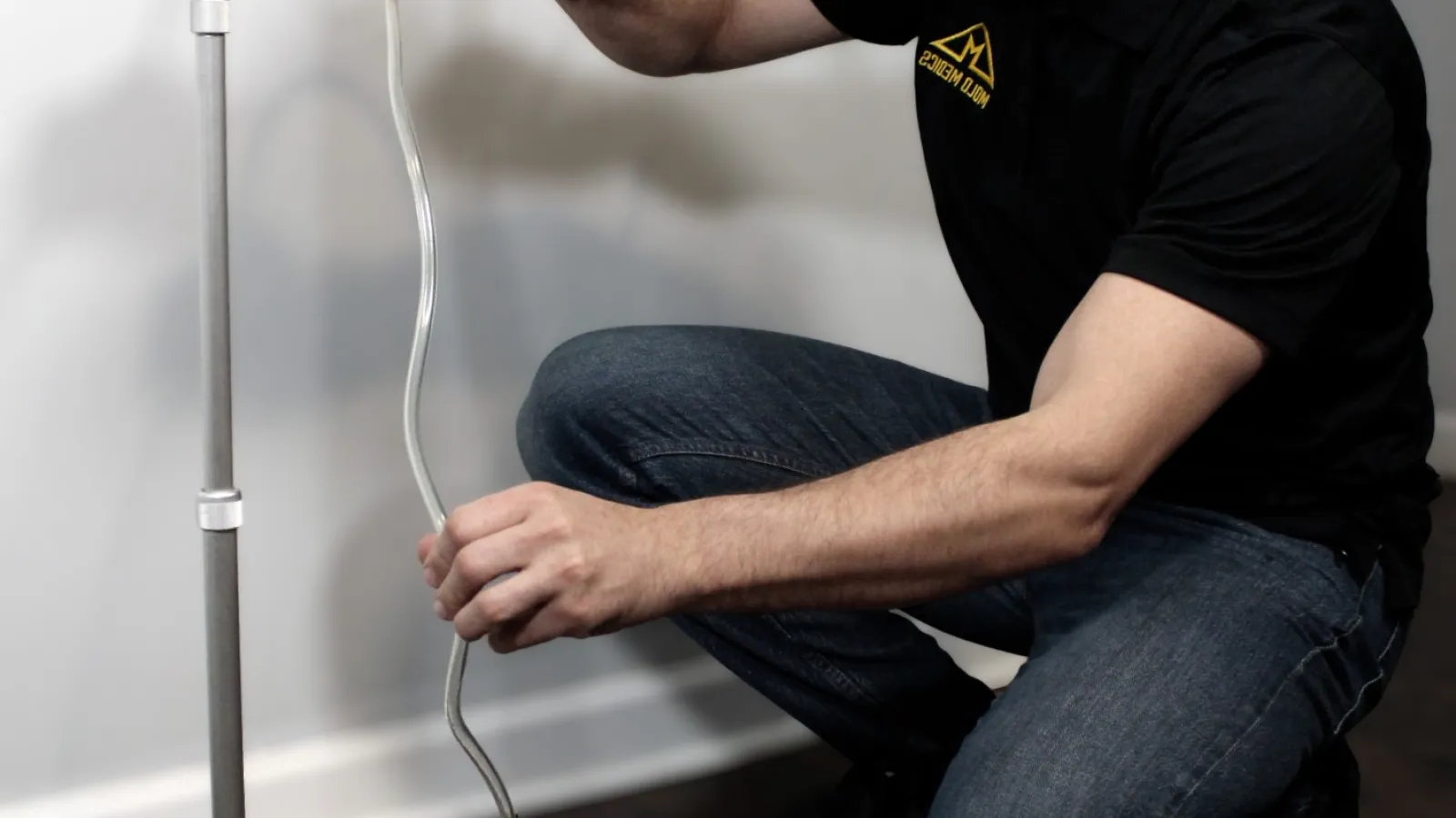 Technician kneeling installing or adjusting a building moisture detection device on hardwood floor near white wall.