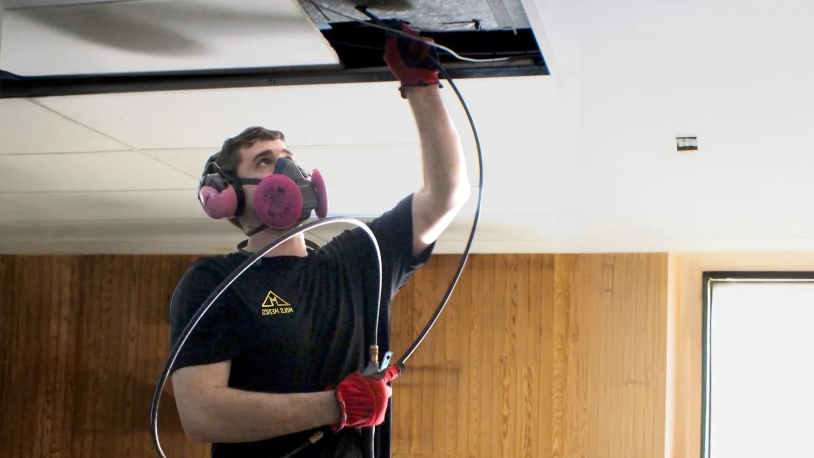 Worker wearing protective mask and gloves repairing ceiling wiring in a modern indoor space.