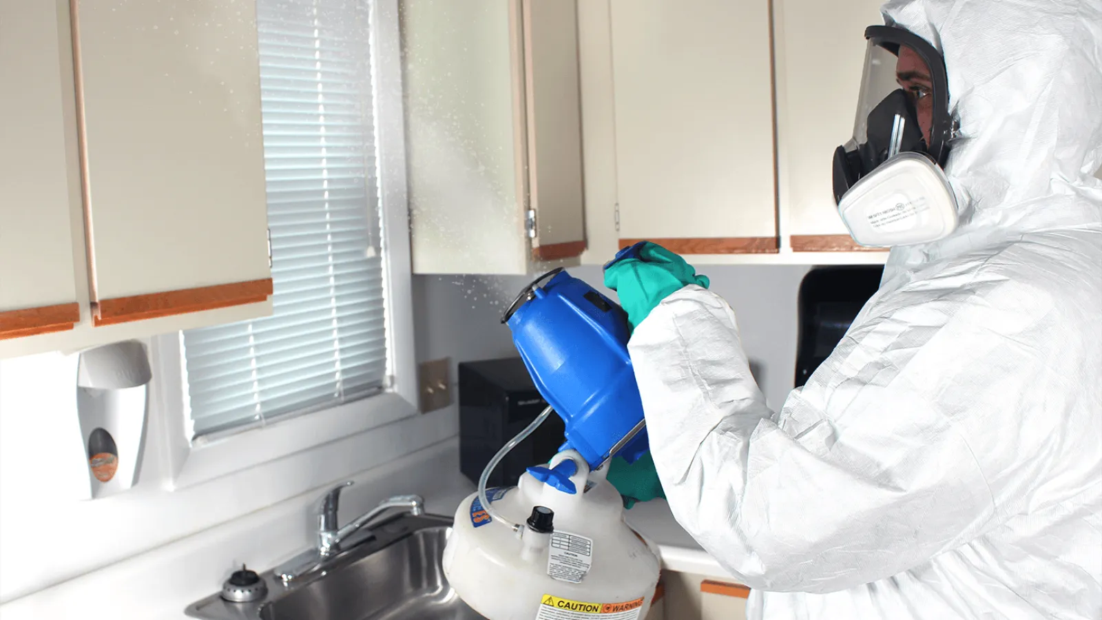 Person in protective suit and mask disinfecting a kitchen area using a fogging device near the sink and cabinets.