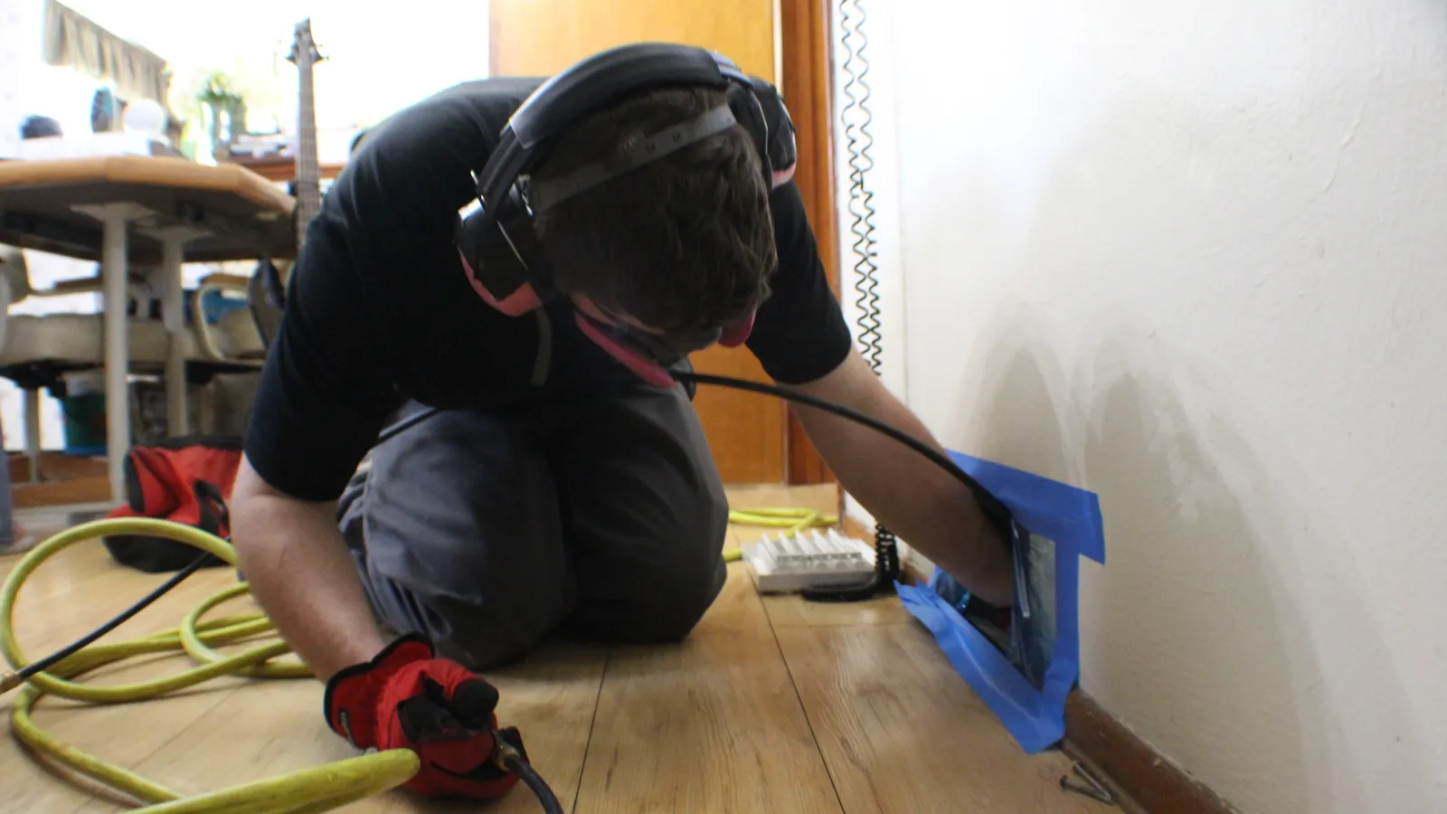 Person wearing protective gear works on a floor outlet installation on wooden floor next to white wall.